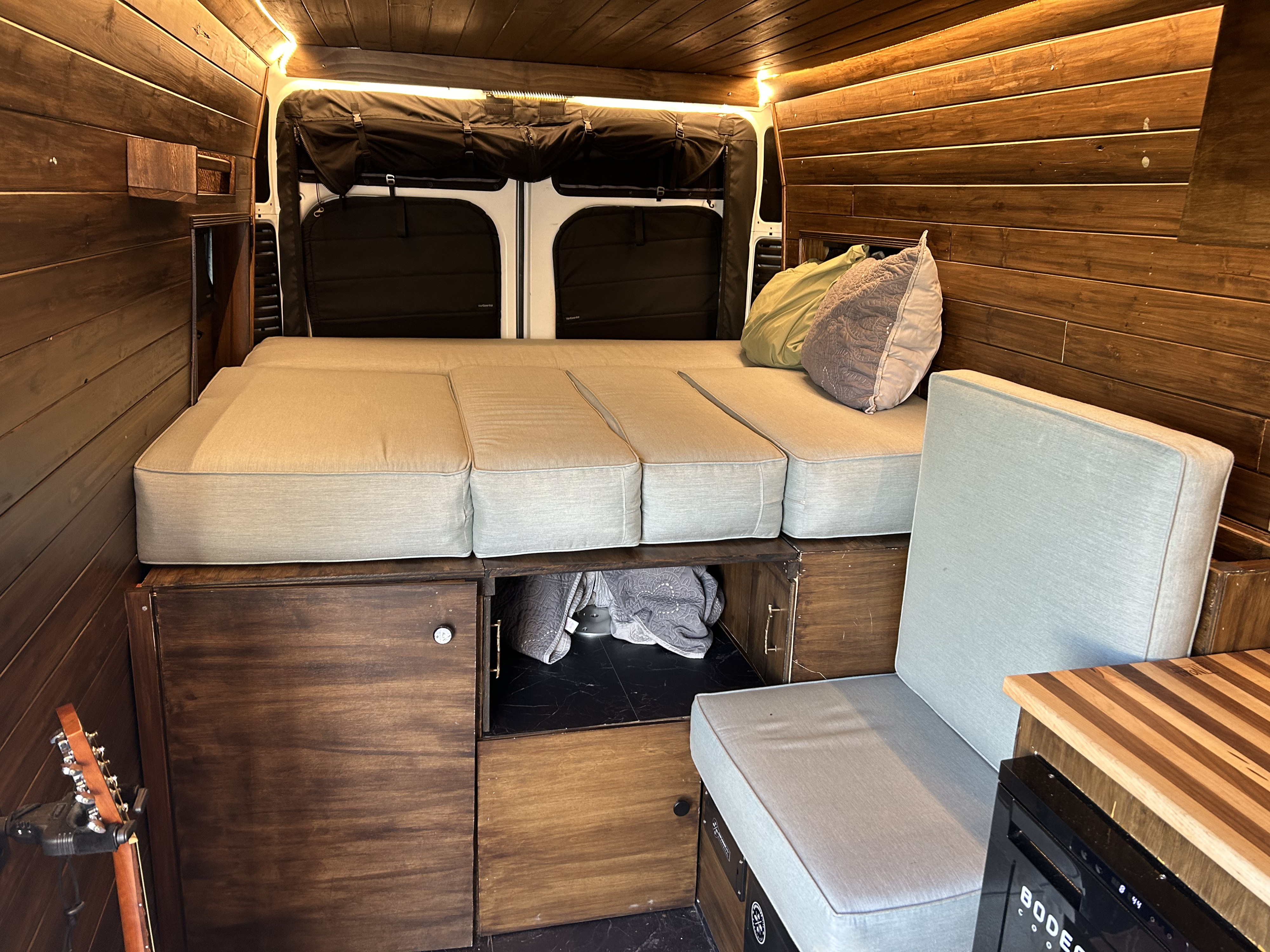 Cozy wood-paneled RV rental interior, featuring a comfortable bed, cushioned seating, built-in storage, and warm lighting.. RAM Promaster 2022