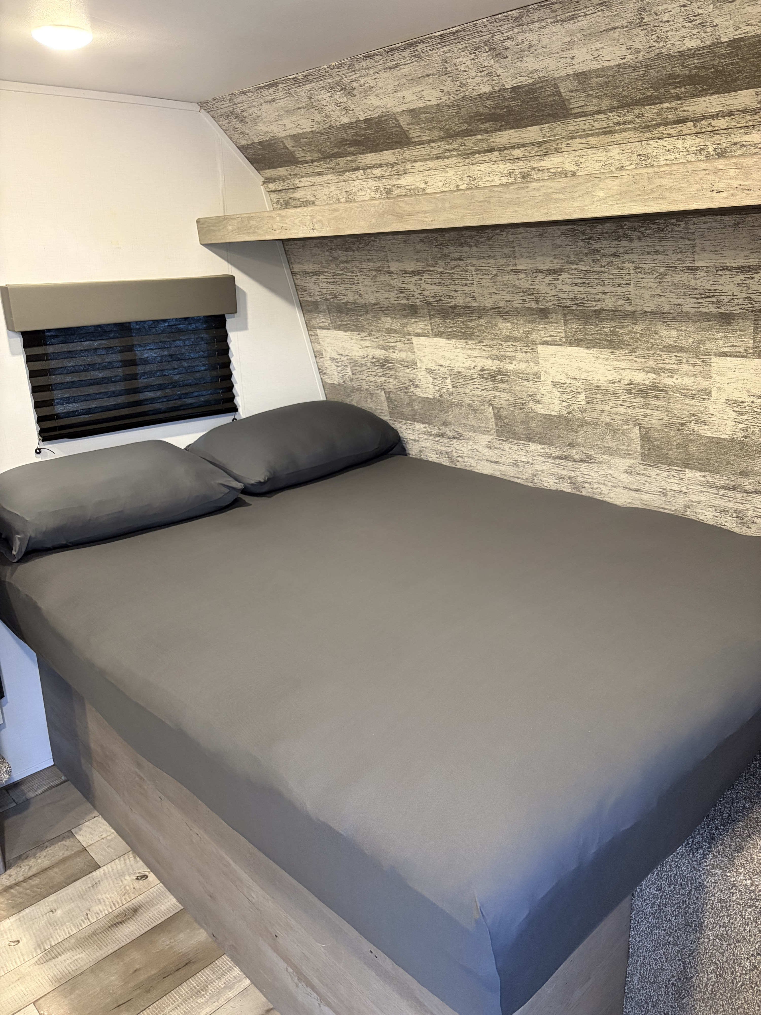 RV rental bedroom with a neatly made bed, grey sheets, pillows, window with blind, and a rustic plank wall.. Keystone RV Springdale 2021