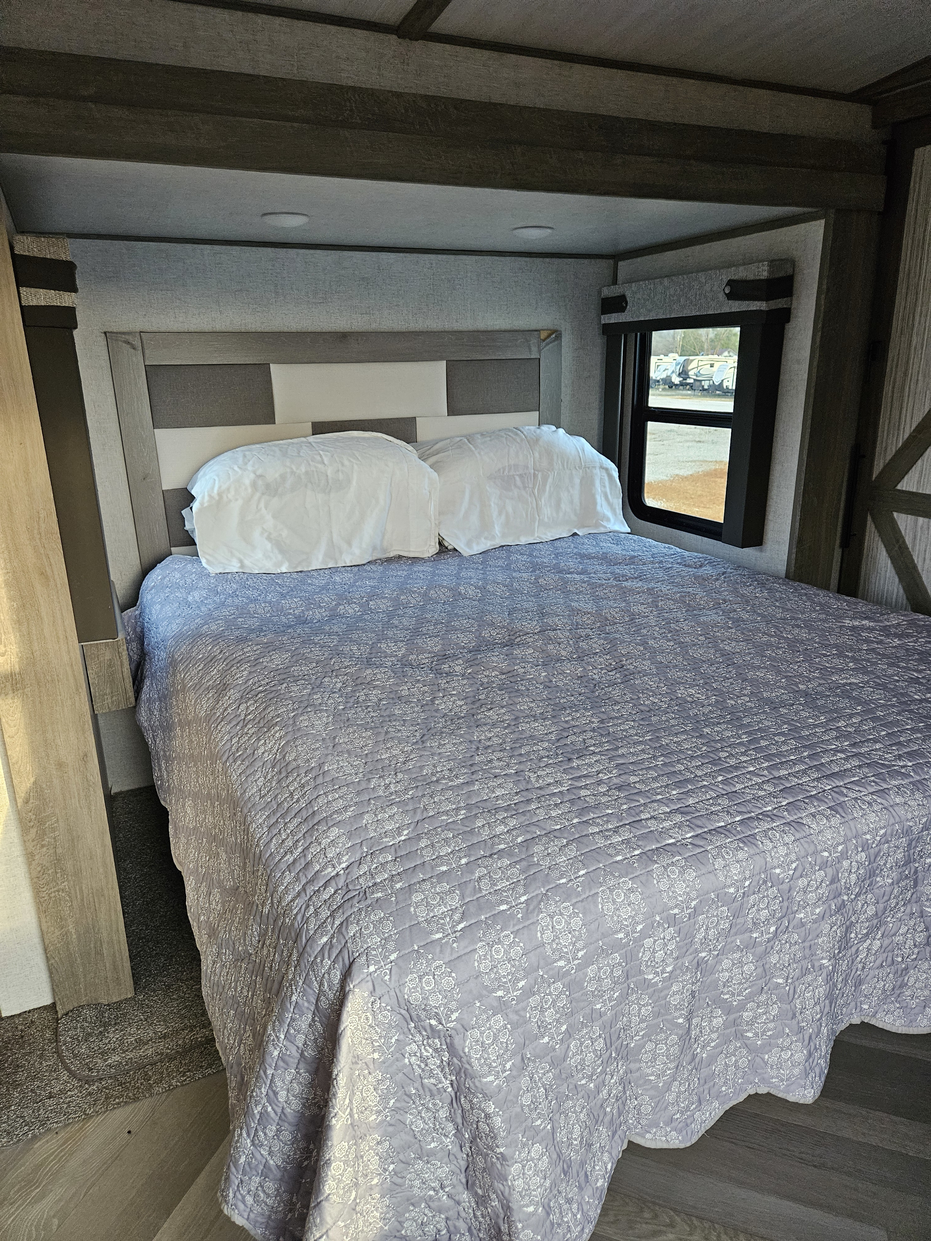 This RV rental bedroom features a cozy bed with a quilted comforter, panel headboard, and a window offering a view outside.. Forest River Wildwood Heritage Glen 2021