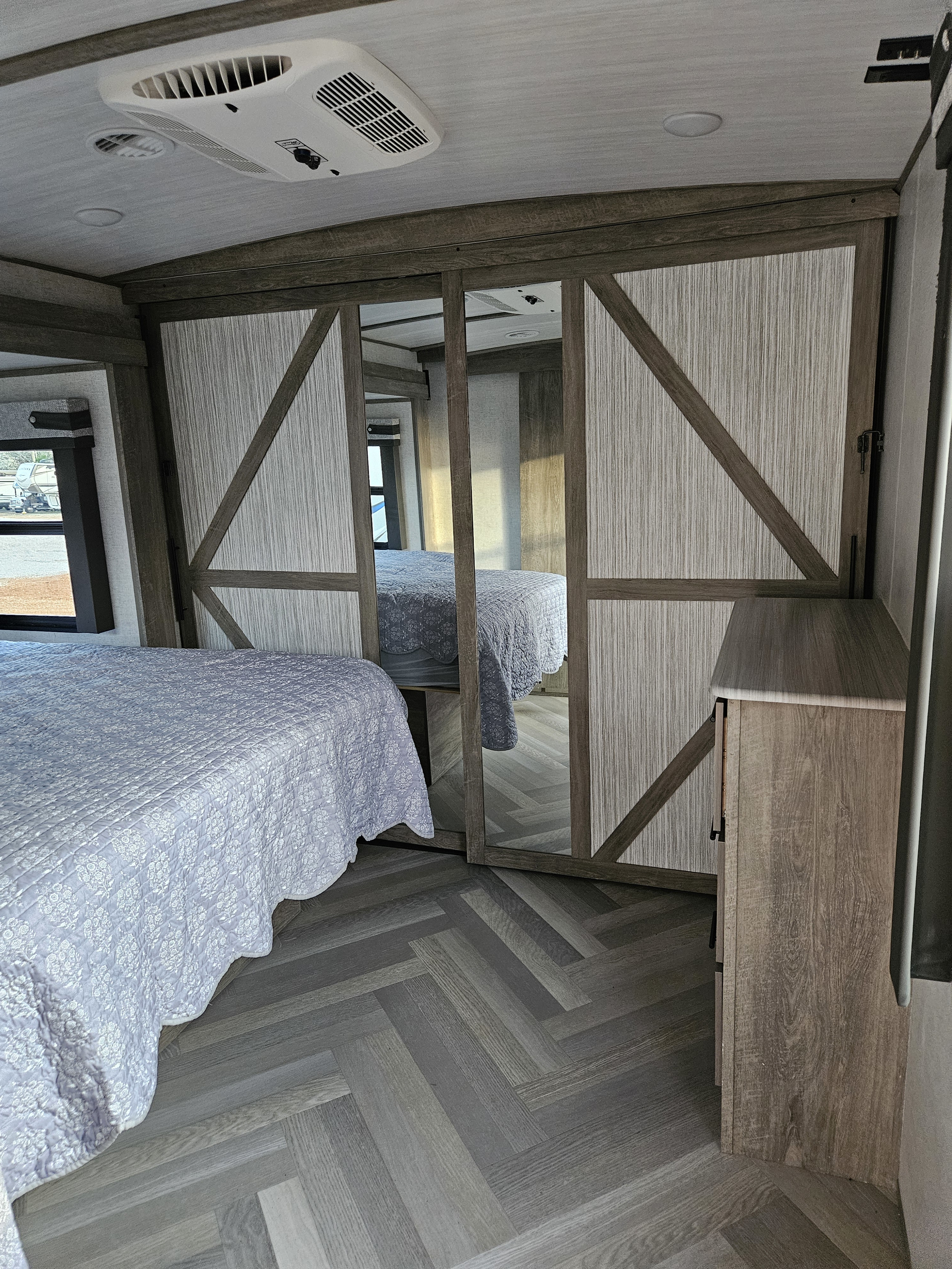 RV rental interior features a cozy bedroom with patterned bedding, a mirrored closet, ceiling AC, and stylish chevron flooring.. Forest River Wildwood Heritage Glen 2021