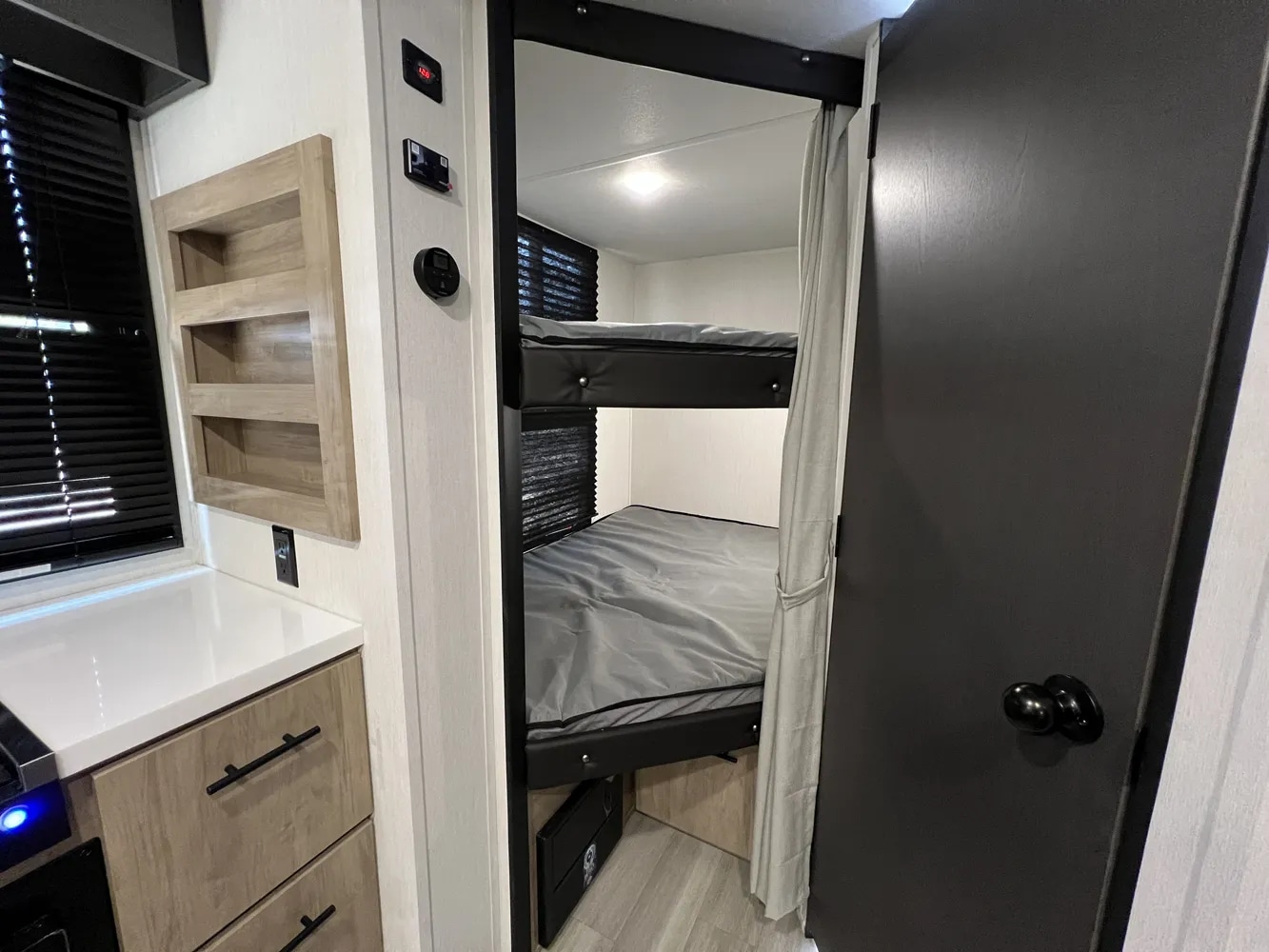 Inside an RV rental, a compact bunk bed area with gray bedding is visible. To the left, a counter, storage, and wall controls are shown.. Forest River Campsite Reserve 2026