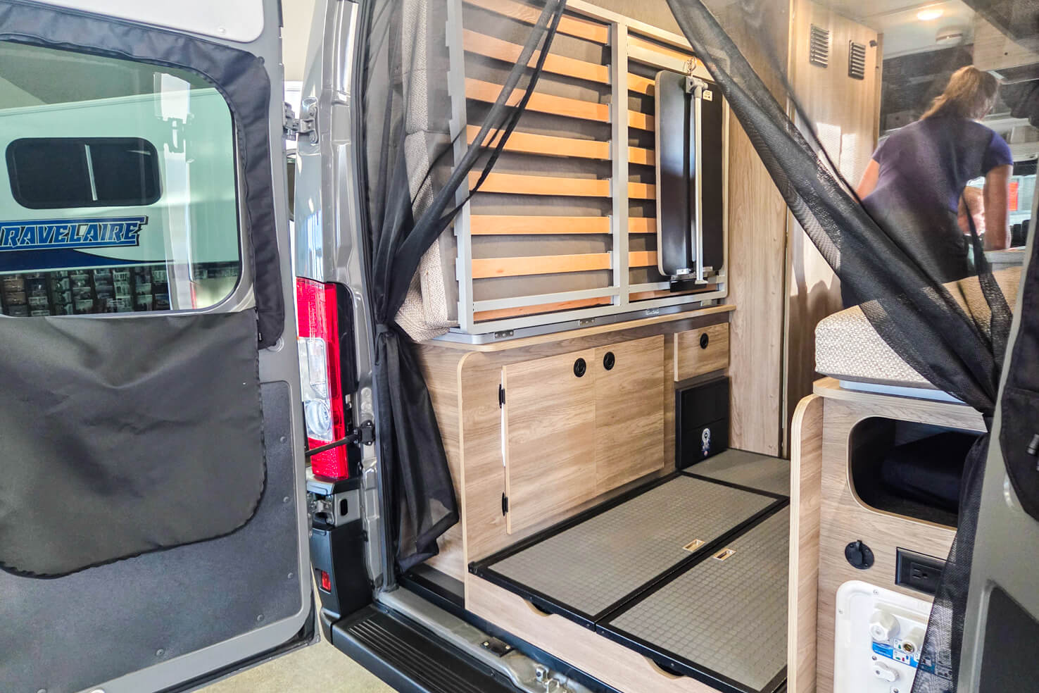 View inside an rv rental camper van from the open rear doors, showing a folded bed frame, wood storage, and black mesh screen.. Winnebago Solis 2025
