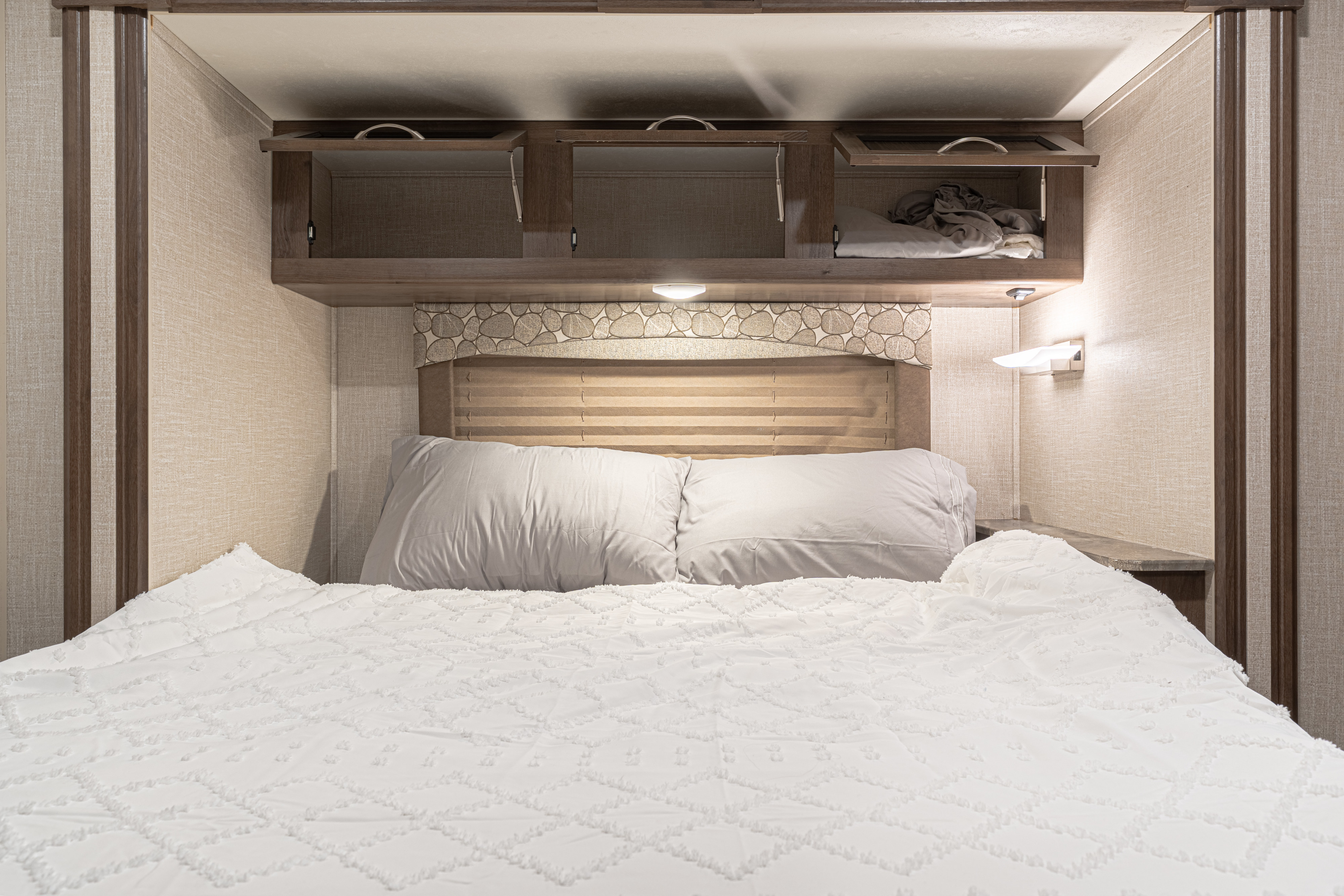Cozy RV rental bedroom featuring a comfortable bed with textured white bedding, overhead storage cabinets, and reading lights.. Forest River Coachmen Freelander 2020