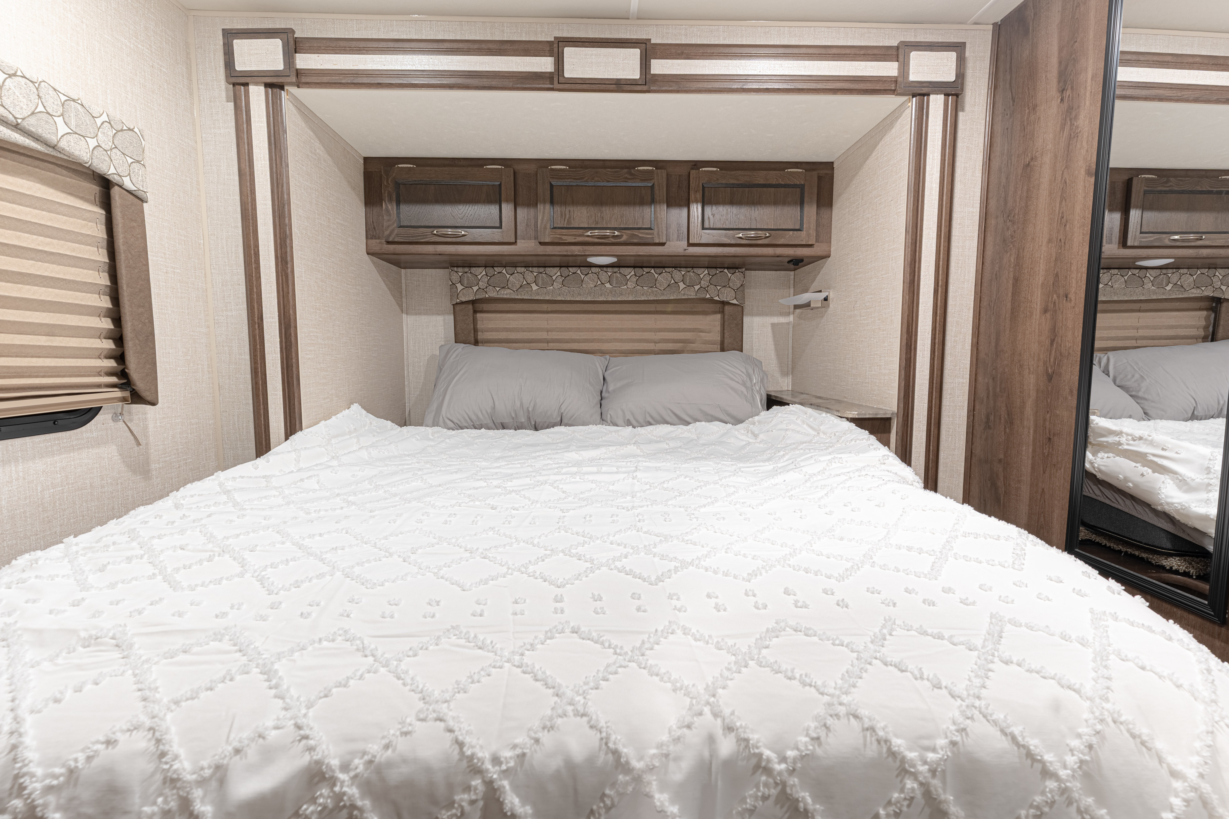 Comfortable RV rental bedroom: white textured bed, gray pillows, wooden overhead cabinets, window, and mirrored closet.. Forest River Coachmen Freelander 2020