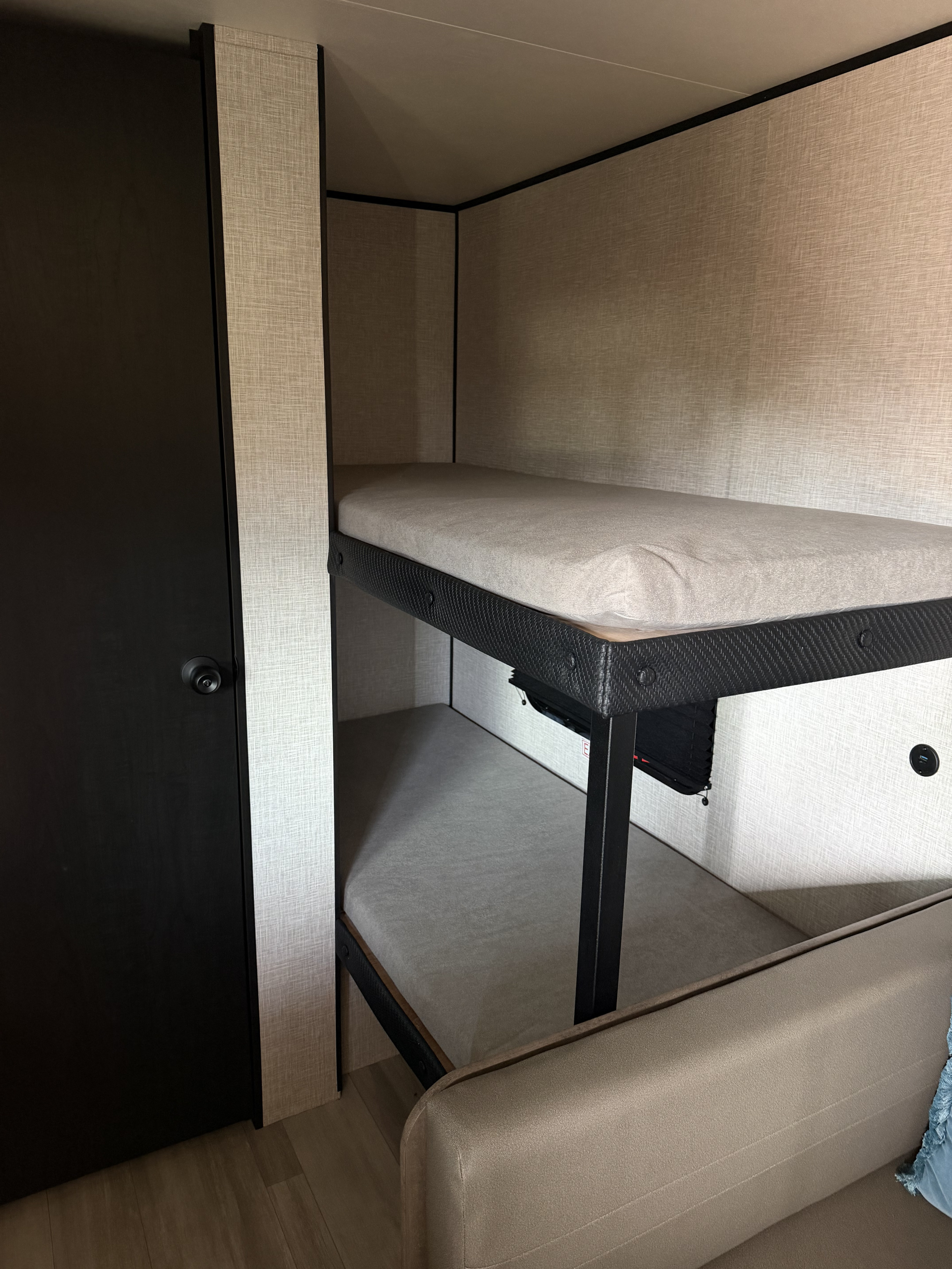 Interior of an rv rental showing compact bunk beds with grey mattresses, textured walls, a black door, and a partial sofa.. Starcraft AR One 2026