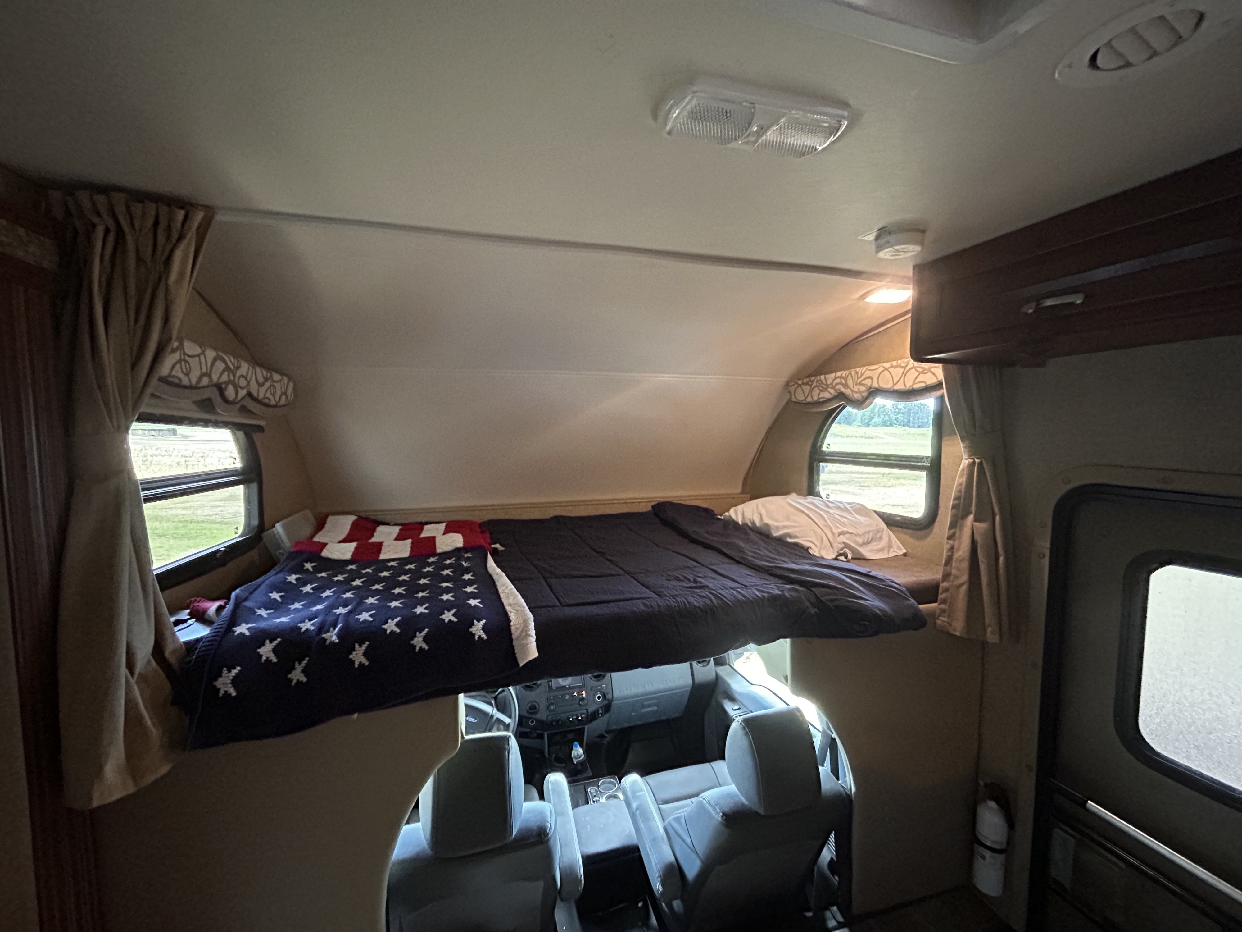 Inside a spacious RV rental: comfortable cab-over bunk with an American flag blanket, overlooking the driver's area.. Thor Four Winds 2015