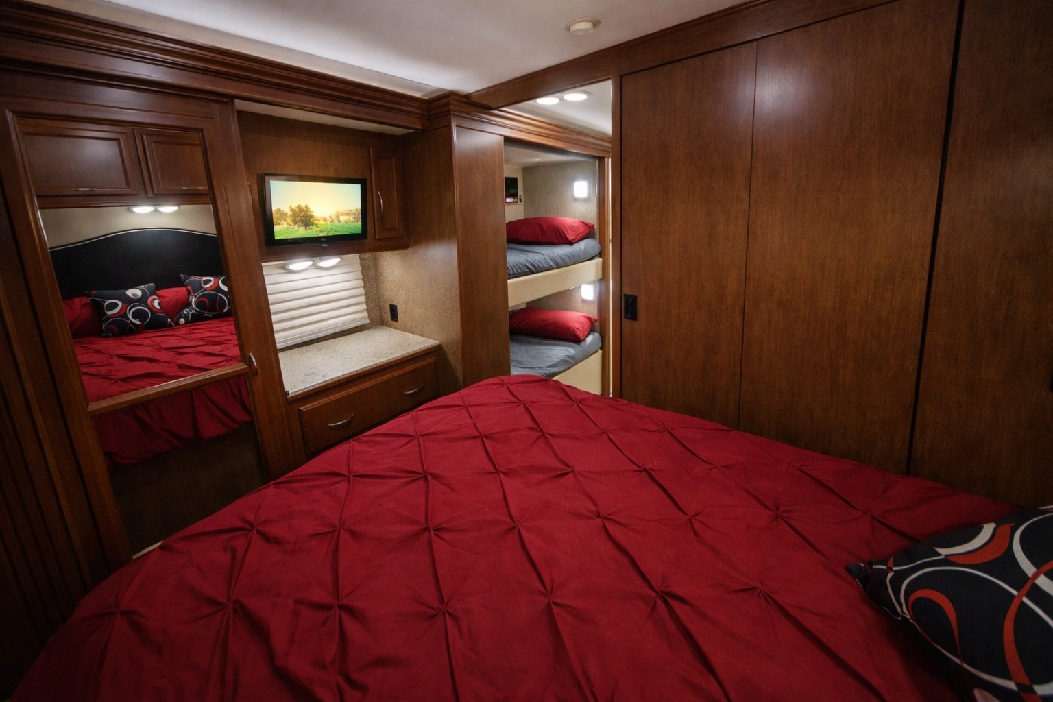 RV rental bedroom featuring a main bed with red bedding, cozy bunk beds, and a mounted TV in a wooden interior.. Thor A.C.E 30.2 2014