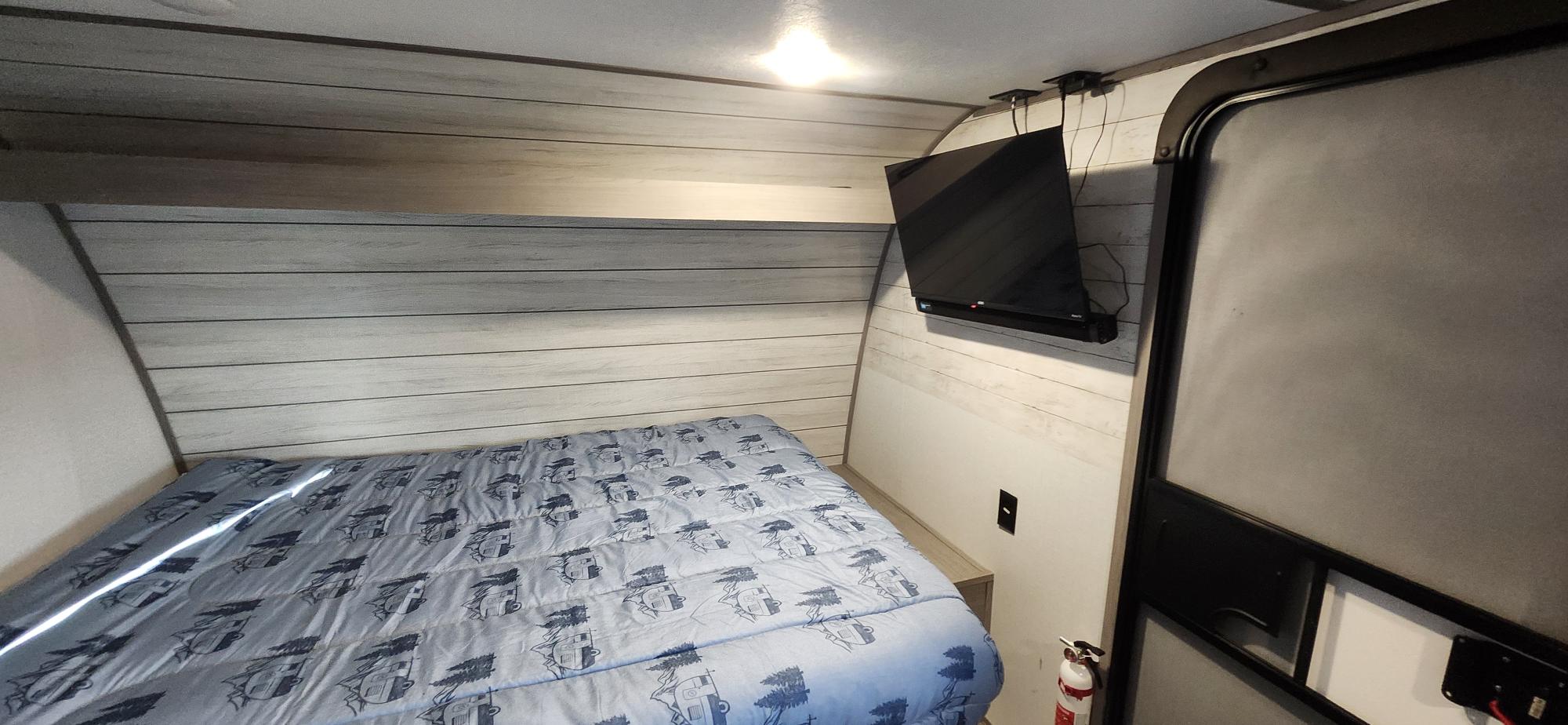 Compact RV rental bedroom with patterned bed, shiplap walls, mounted TV, and a fire extinguisher by the door.. Dutchmen Coleman Lantern LT 2023