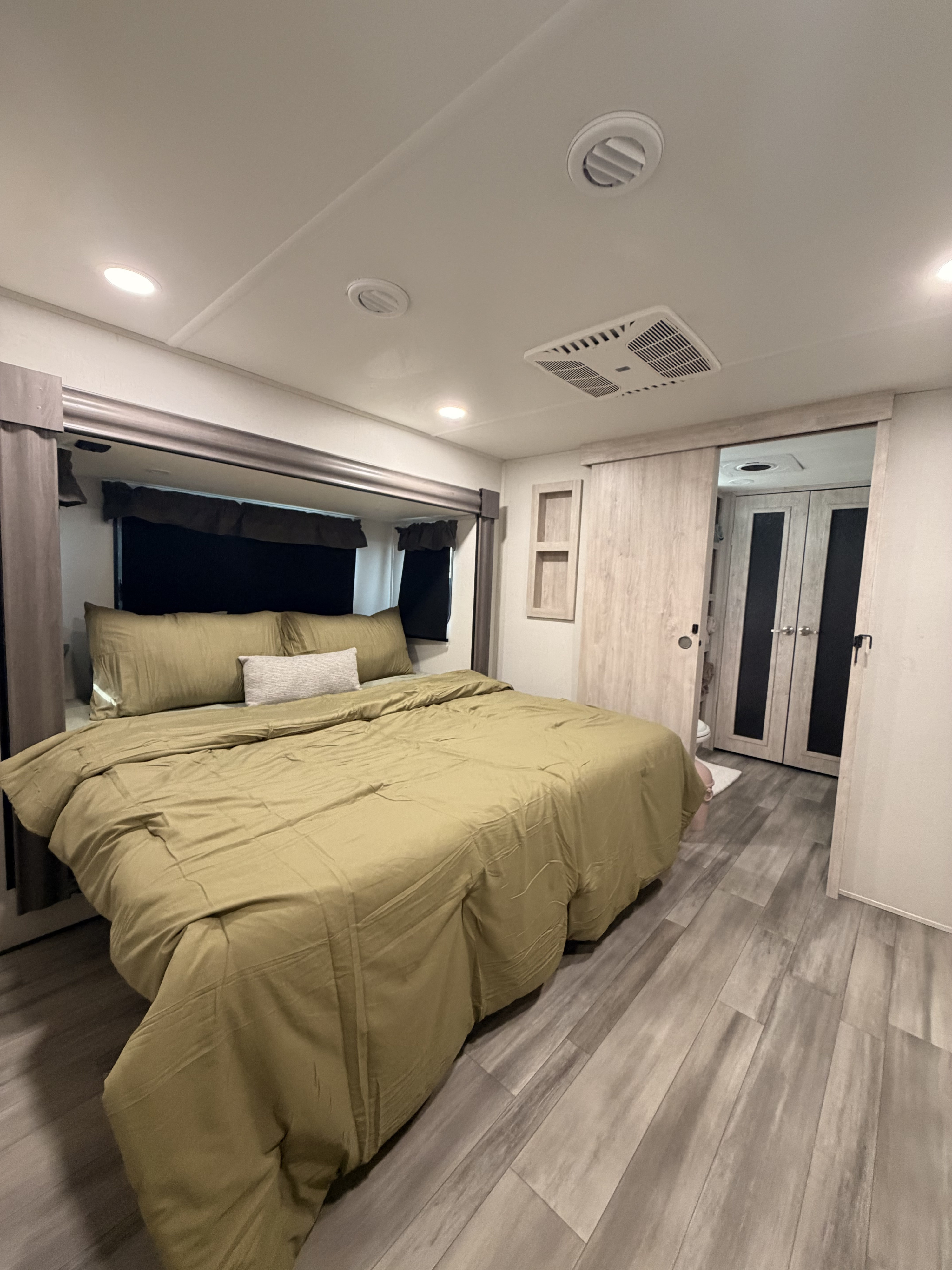Cozy RV rental bedroom with olive green bedding, modern lighting & AC. Light wood-look flooring and a sliding door. Ideal for travel.. Forest River Sierra 2024