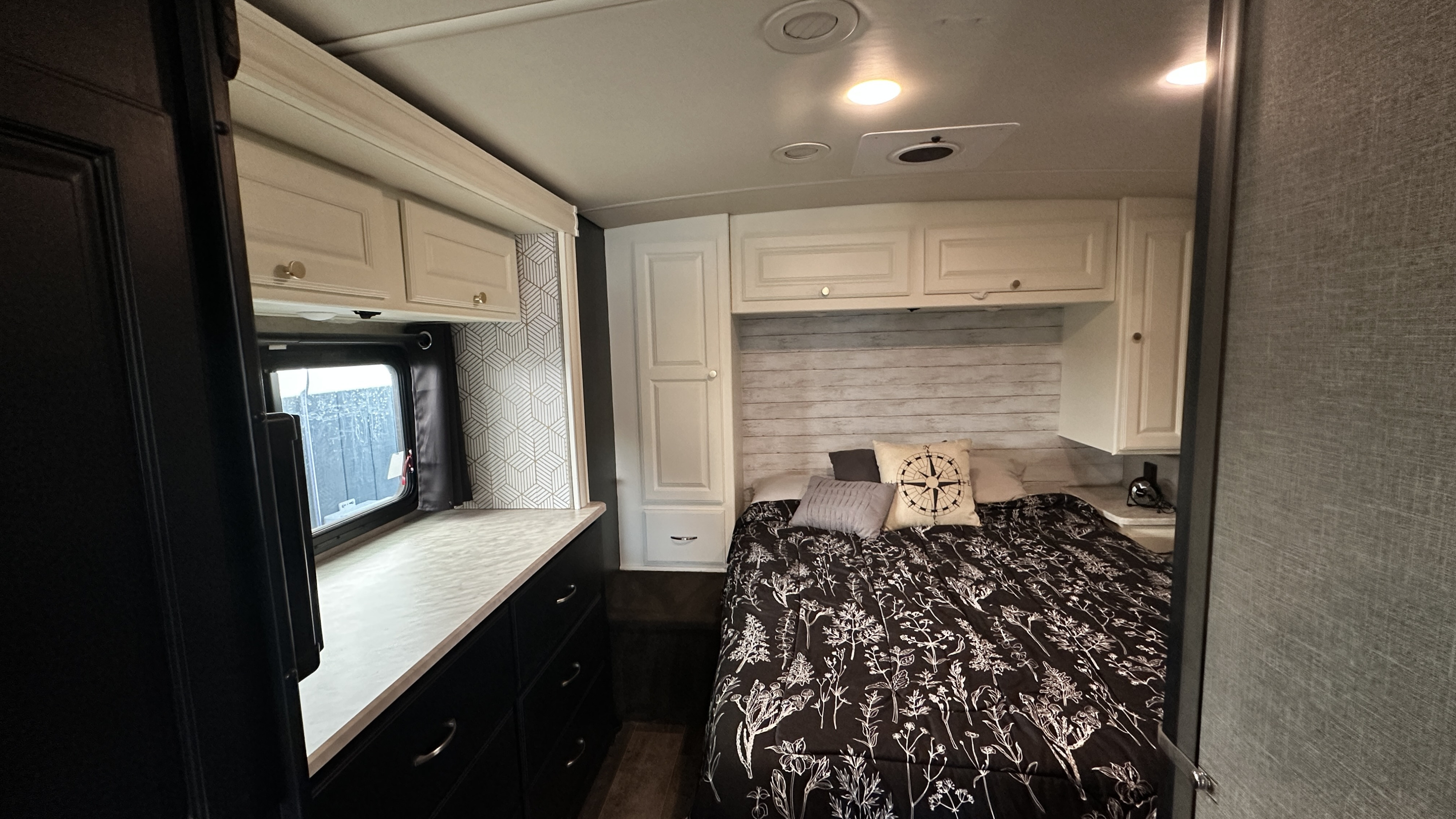 Cozy RV rental bedroom featuring a bed with botanical bedding, decorative pillows, white overhead cabinets, dark lower drawers, and patterned walls.. Winnebago Vista LX 2019