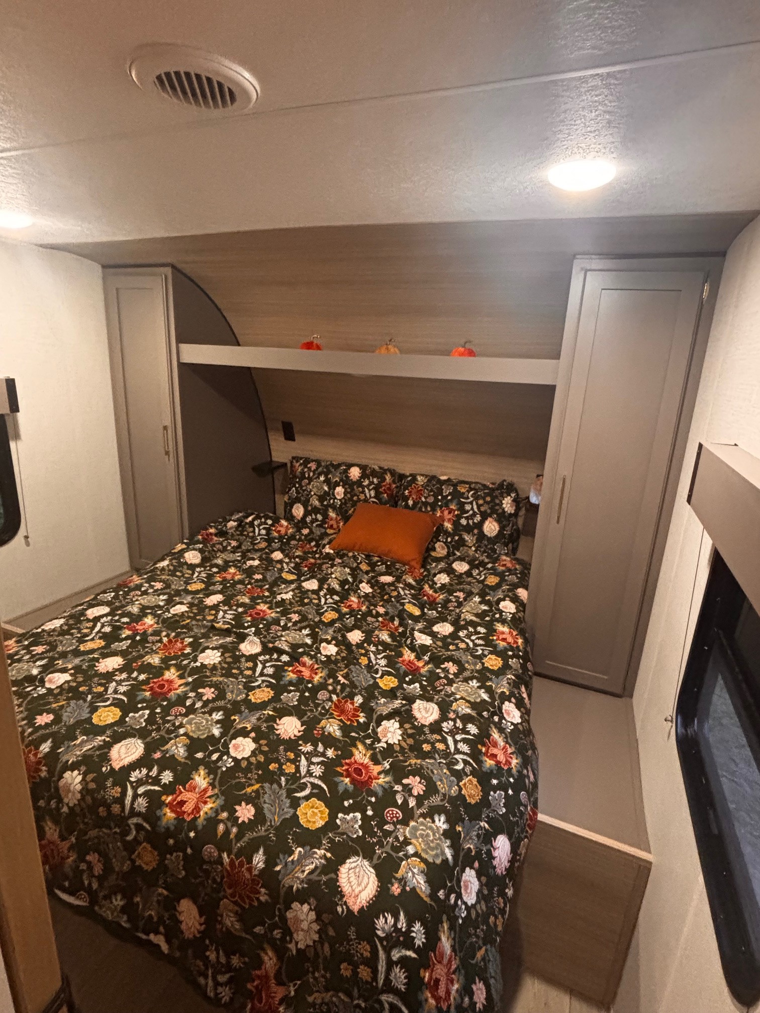 Cozy RV rental bedroom with a vibrant floral comforter, orange accent pillow, overhead cabinets, and warm lighting.. KZ Sportsmen Select 2025