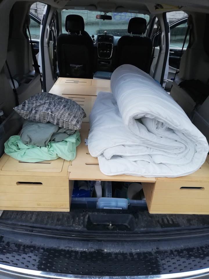RV rental van interior with a cozy bed setup for camping, featuring bedding, pillows, and under-platform storage for a road trip.. Dodge Grand Caravan 2012