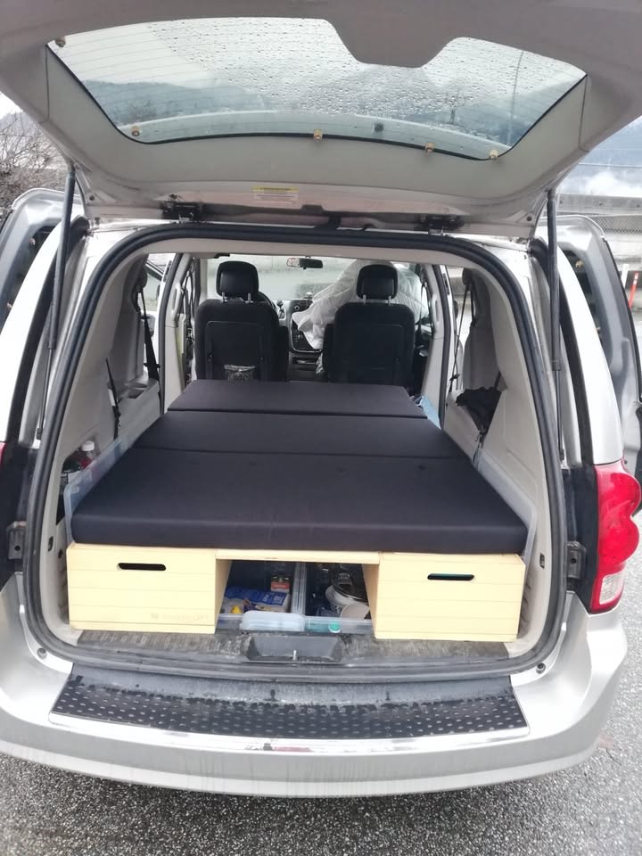 Inside a minivan, a DIY wooden bed platform with a black mattress and storage offers an RV rental alternative for car camping.. Dodge Grand Caravan 2012