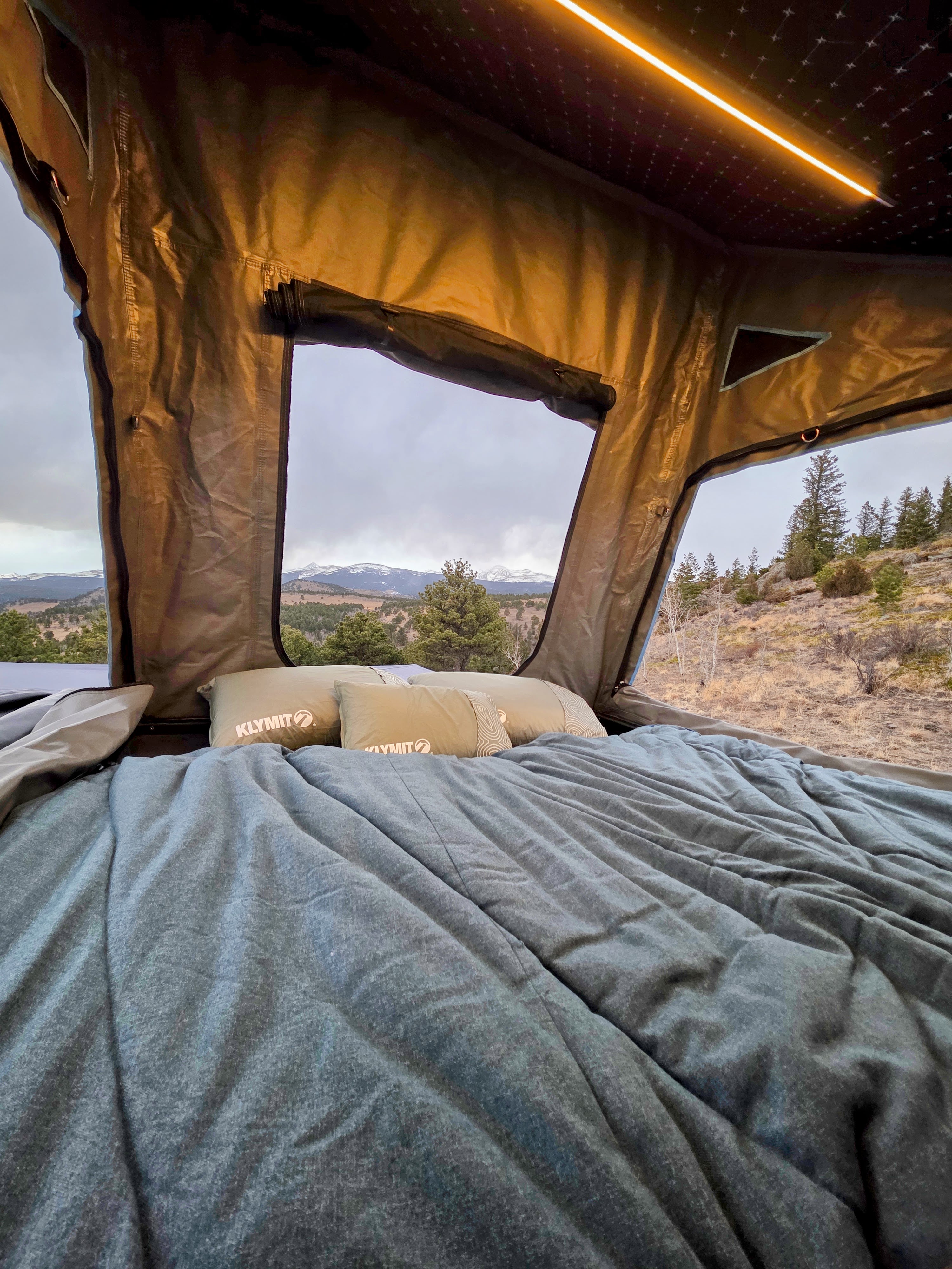Inside a comfy RV rental rooftop tent, showcasing Klymit pillows, a cozy bed, and stunning mountain views under a cloudy sky.. Dirtbox Overland Canopy Camper 2023