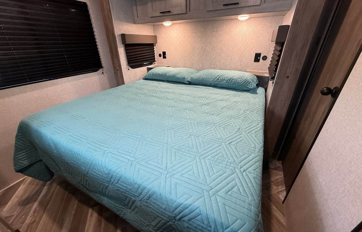 RV rental bedroom: a comfy bed with a blue quilt, pillows, window blinds, and overhead cabinets.. Crossroads RV Zinger 2025