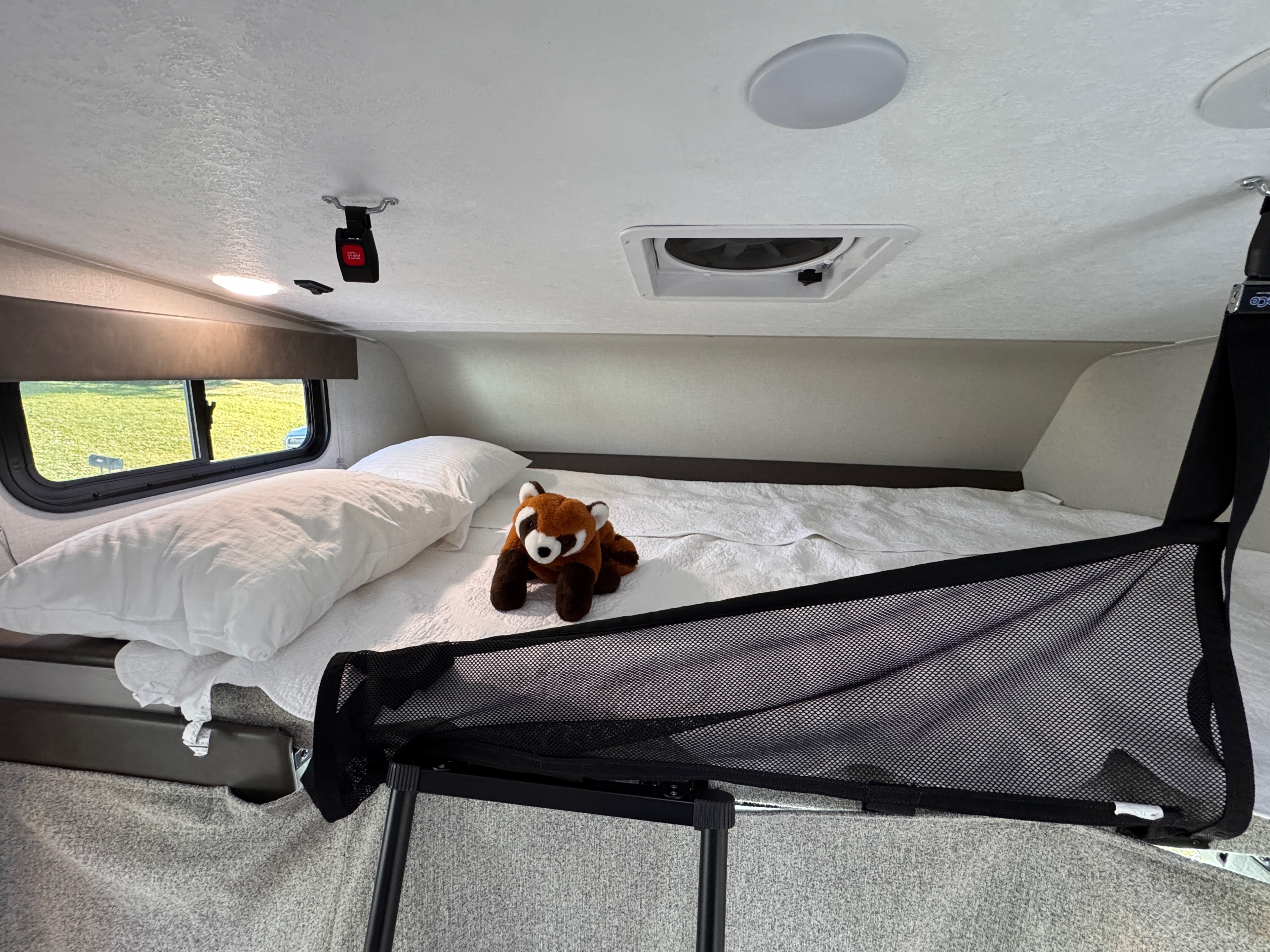 Inside an RV rental, this cozy bunk features a bed with white linen, a red panda, safety net, and a window view.. Forest River Coachmen Freelander 2025