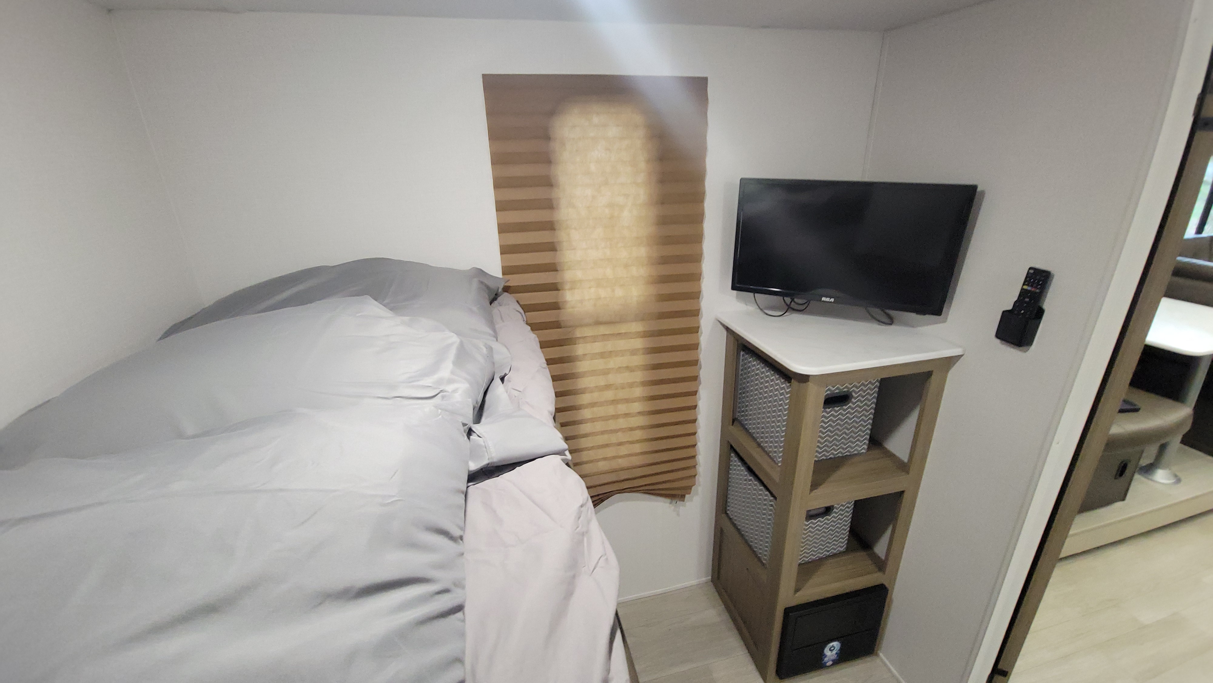 An RV rental bedroom with a neatly made bed, TV, storage shelf unit with grey bins, and a pleated window blind.. Forest River Salem Cruise-Lite 2025