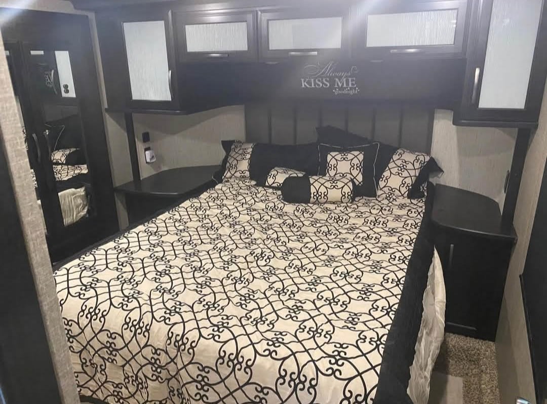 Inside a modern RV rental: a comfortable bedroom with a large bed, elegant black & white bedding, overhead storage, and a mirrored wardrobe.. Grand Design Momentum 2019