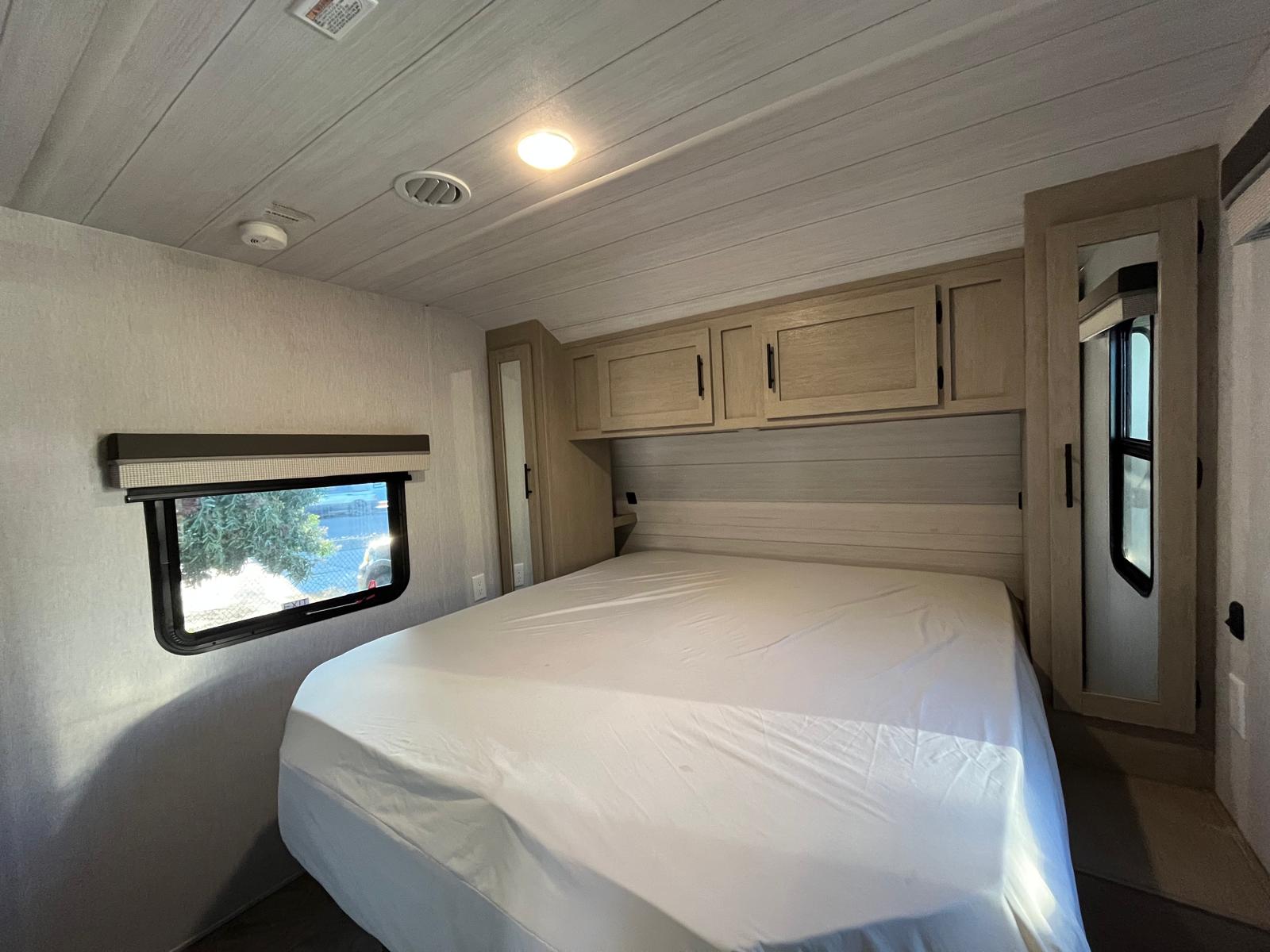 RV rental bedroom with bed, white sheets, wood cabinets, and window. Ceiling light, AC vent, and smoke detector visible.. Forest River Salem 2023