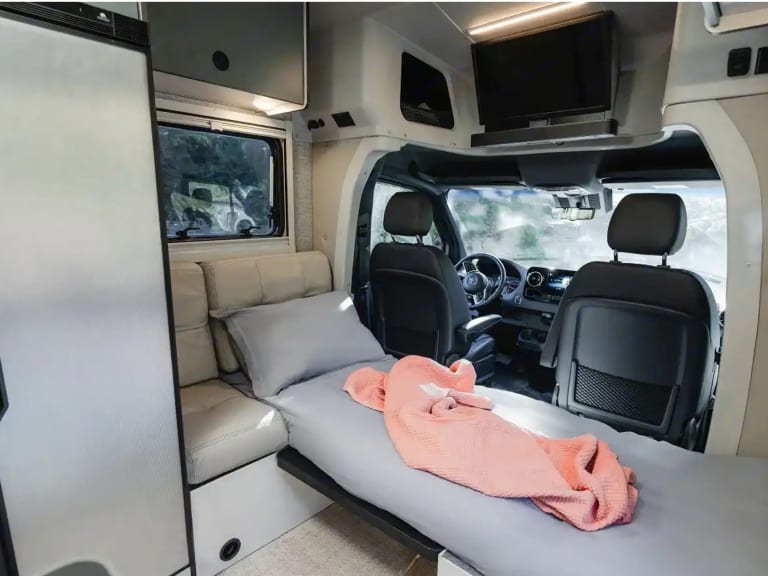 Cozy rv rental interior: Bed with pink blanket, integrated fridge, and front driving cockpit are visible in this campervan.. Winnebago Ekko 2025