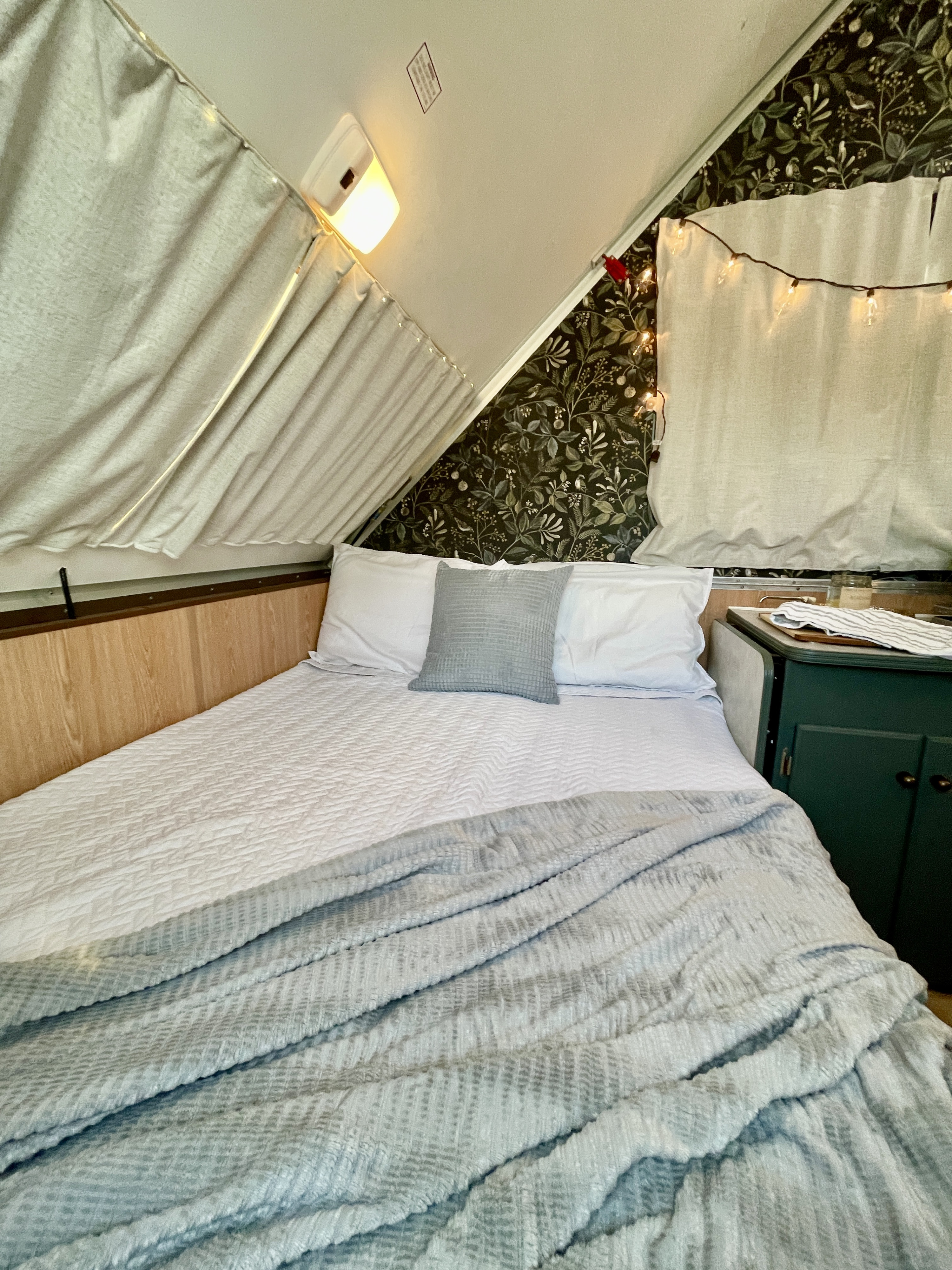 Cozy rv rental bedroom with a made bed, patterned wallpaper, ambient lights, and a light blue blanket.. A Liner A Liner Popup Trailer 2000