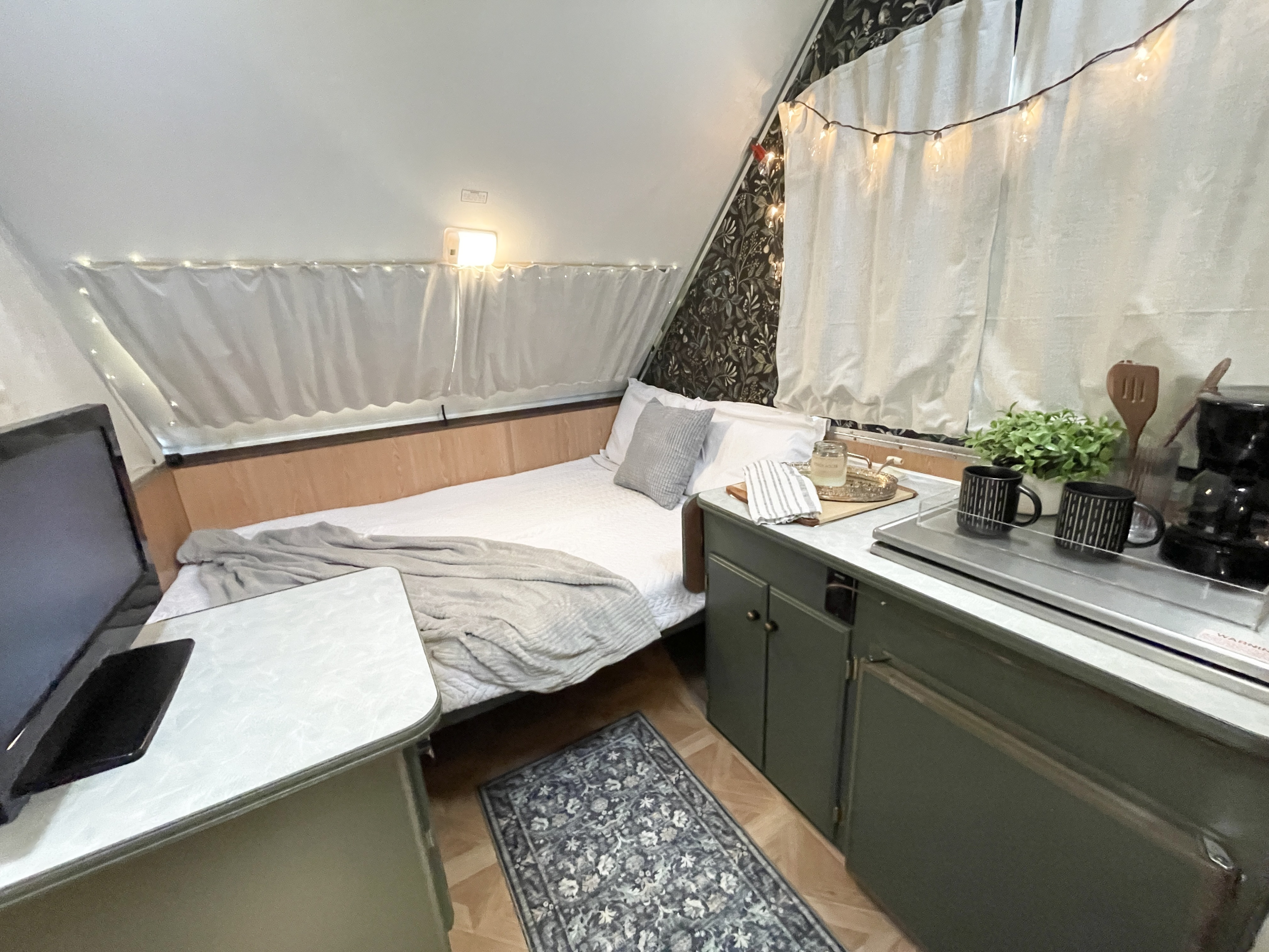 Cozy RV rental interior with a comfortable bed, compact kitchen area, small table with a monitor, and string lights.. A Liner A Liner Popup Trailer 2000