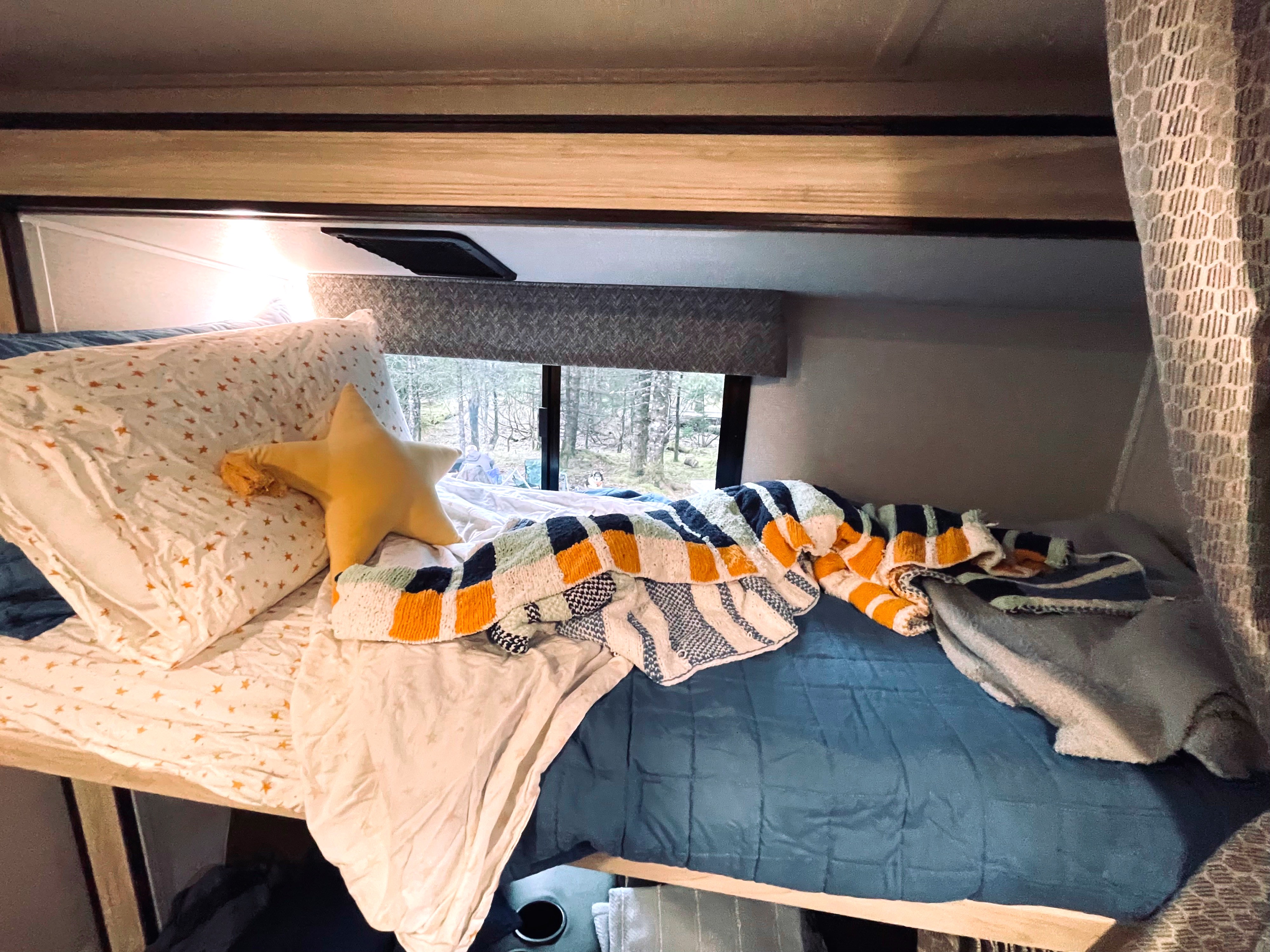 Inside a comfy RV rental: a bunk bed with star pillow & knitted blanket, overlooking a wooded view.. Forester LE 2022