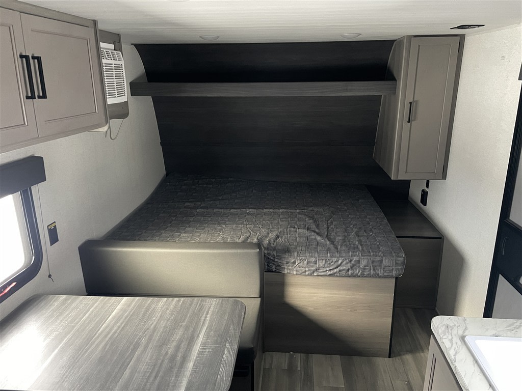 An rv rental interior featuring a bed with overhead storage, a dinette, and a small AC unit. Clean, modern space for travel.. Keystone RV Hideout Sport 2024