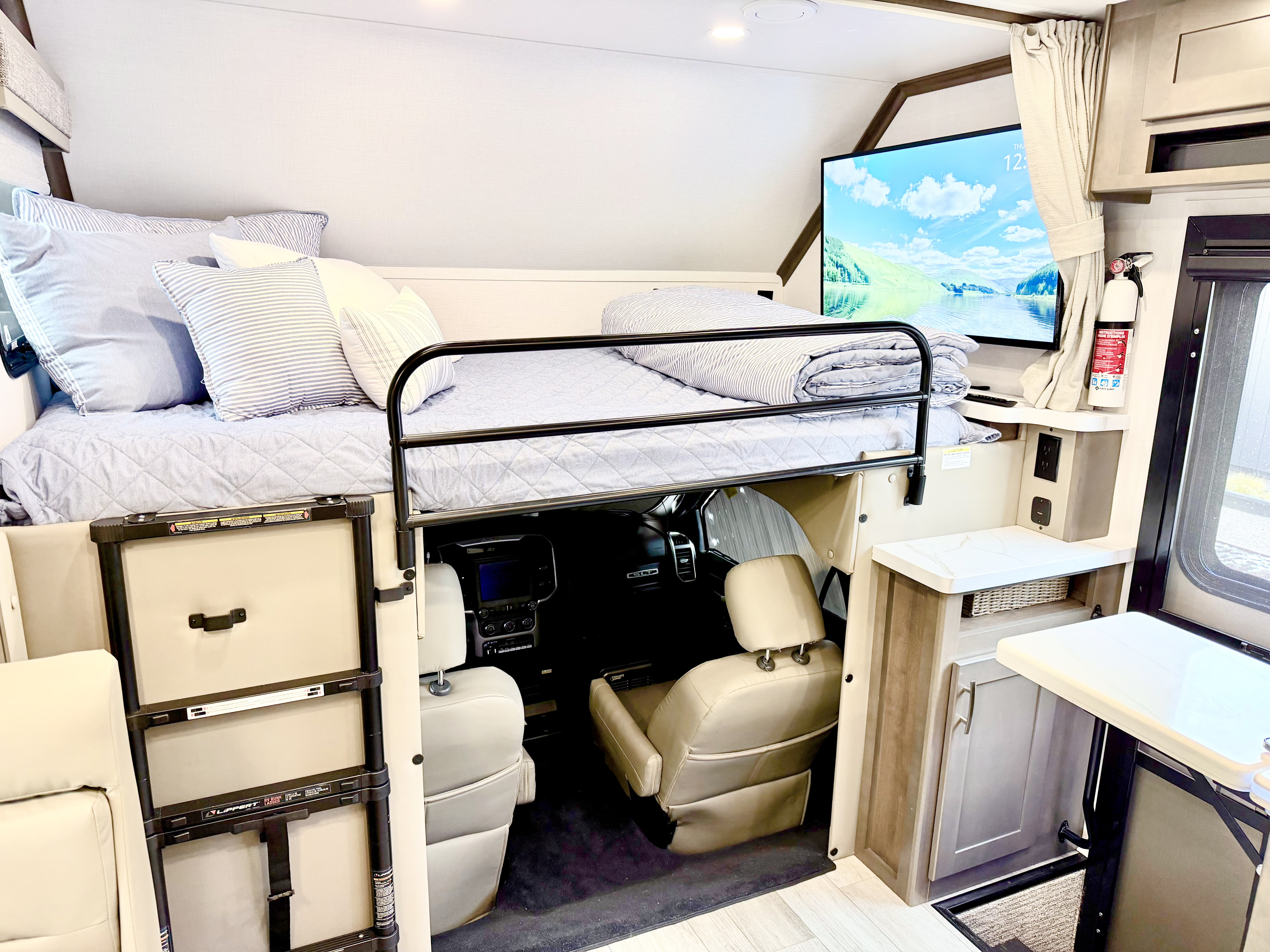 Cozy RV rental cab-over bed with safety rail & TV. Driver's cabin below. Fire extinguisher & storage are visible.. Dynamax Explore ISATA 5 Explorer Extreme Package 2026