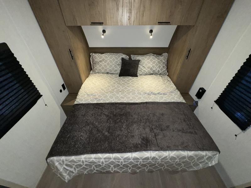 Cozy RV rental bedroom with a queen bed, geometric bedding, overhead storage, and reading lights. Black blinds on angled side walls.. Forest River Cherokee Grey Wolf Black Label 2024