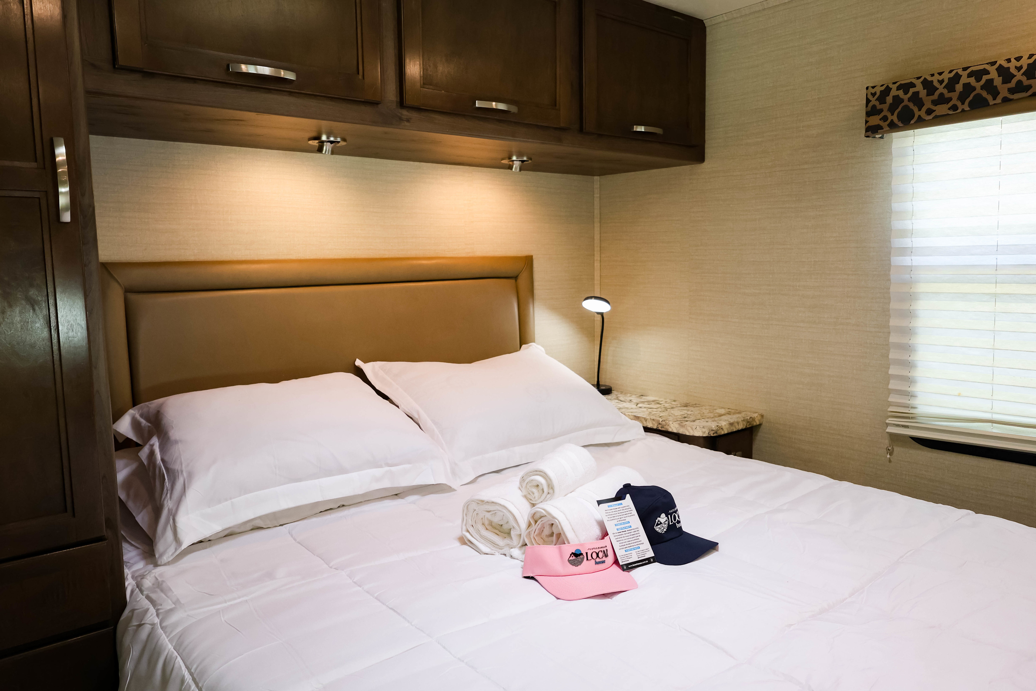 Cozy RV rental bedroom: bed with white linens, overhead cabinets, nightstand with lamp, and window. Welcome hats & rolled towels.. Adventurer Jayco Redhawk 29XK ano 2019 2019
