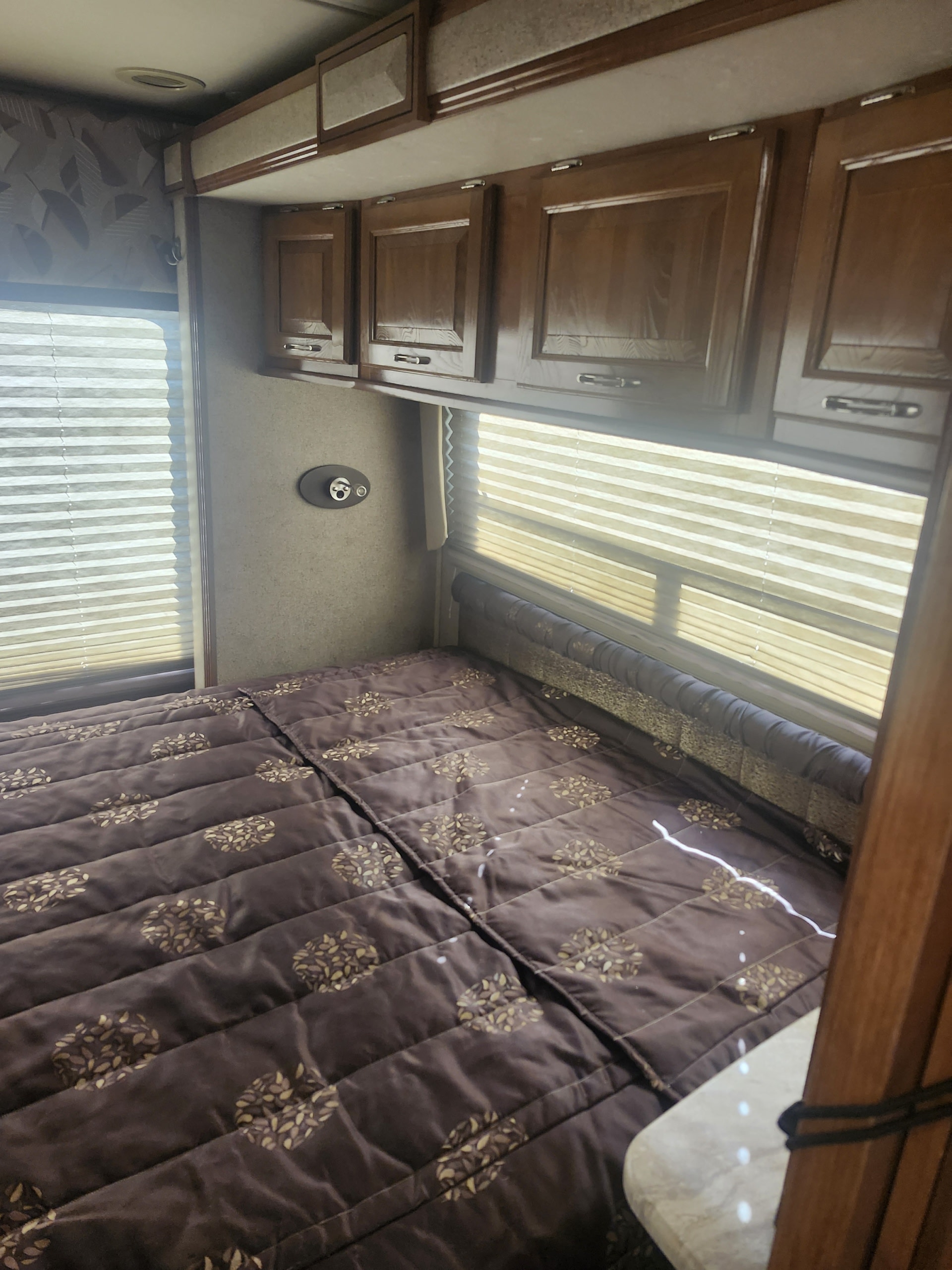 Inside an **rv rental**, a brown quilted bed fills the frame, flanked by windows with blinds and overhead wood cabinets.. Coachman 300 TS 2018