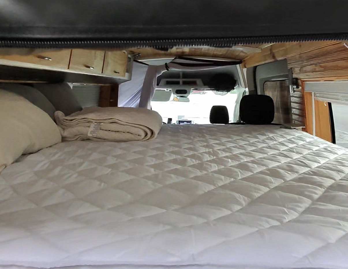 Inside an RV rental: a plush bed with a white quilted mattress pad & pillows. View to front seats & dashboard, cozy wood interior.. Ford Transit 2017