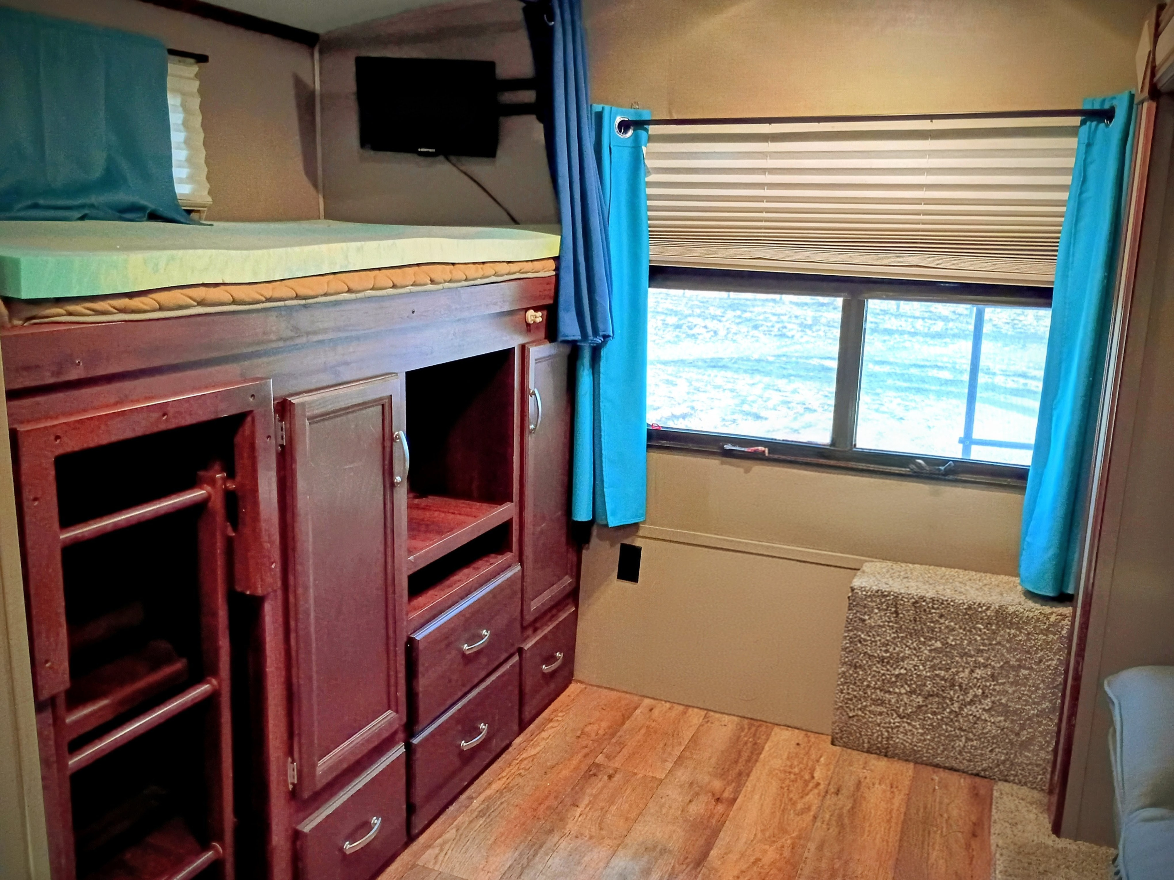 RV rental bunk area: a green foam mattress rests over wooden storage cabinets & drawers, near a window with blinds & a TV.. Keystone RV Outback Super-Lite 2015