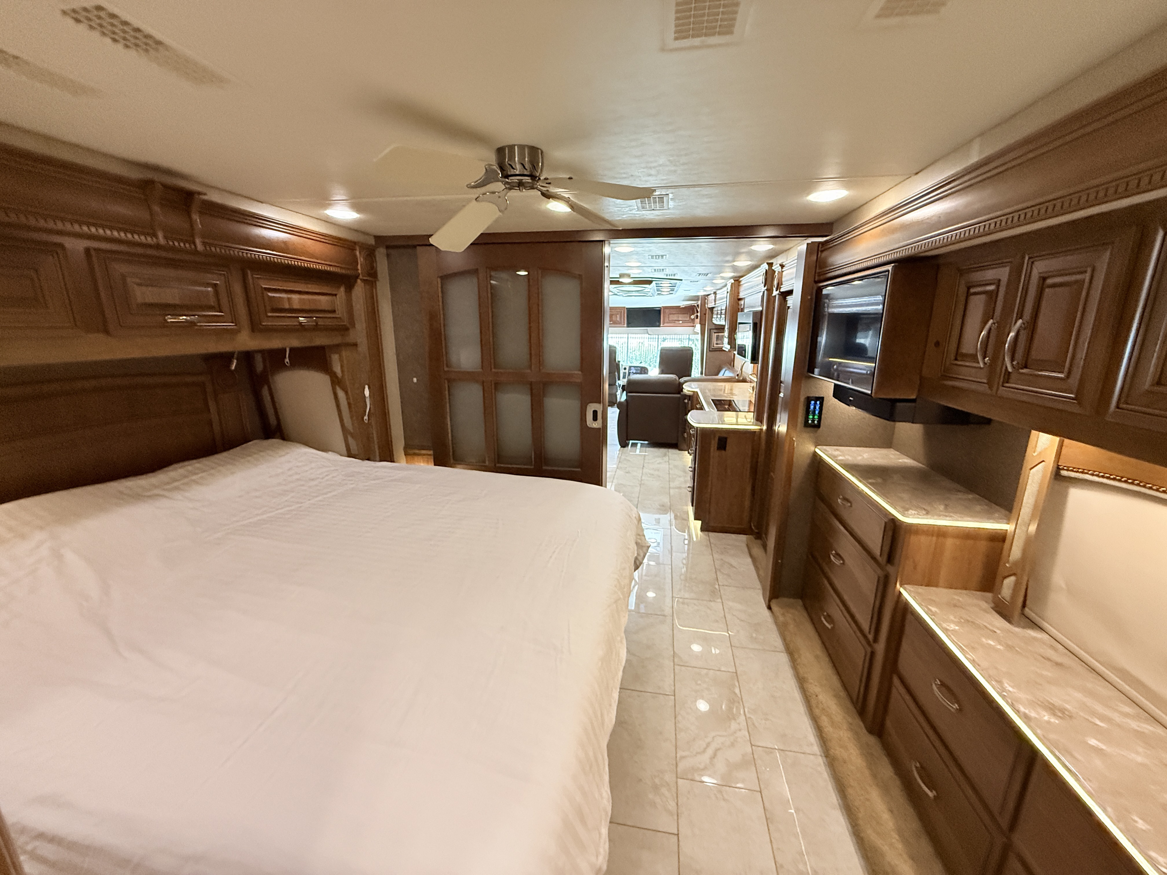 Spacious luxury RV rental bedroom with white bedding, wood cabinetry, a TV, and view into the living area.. Entegra Coach Anthem 2019