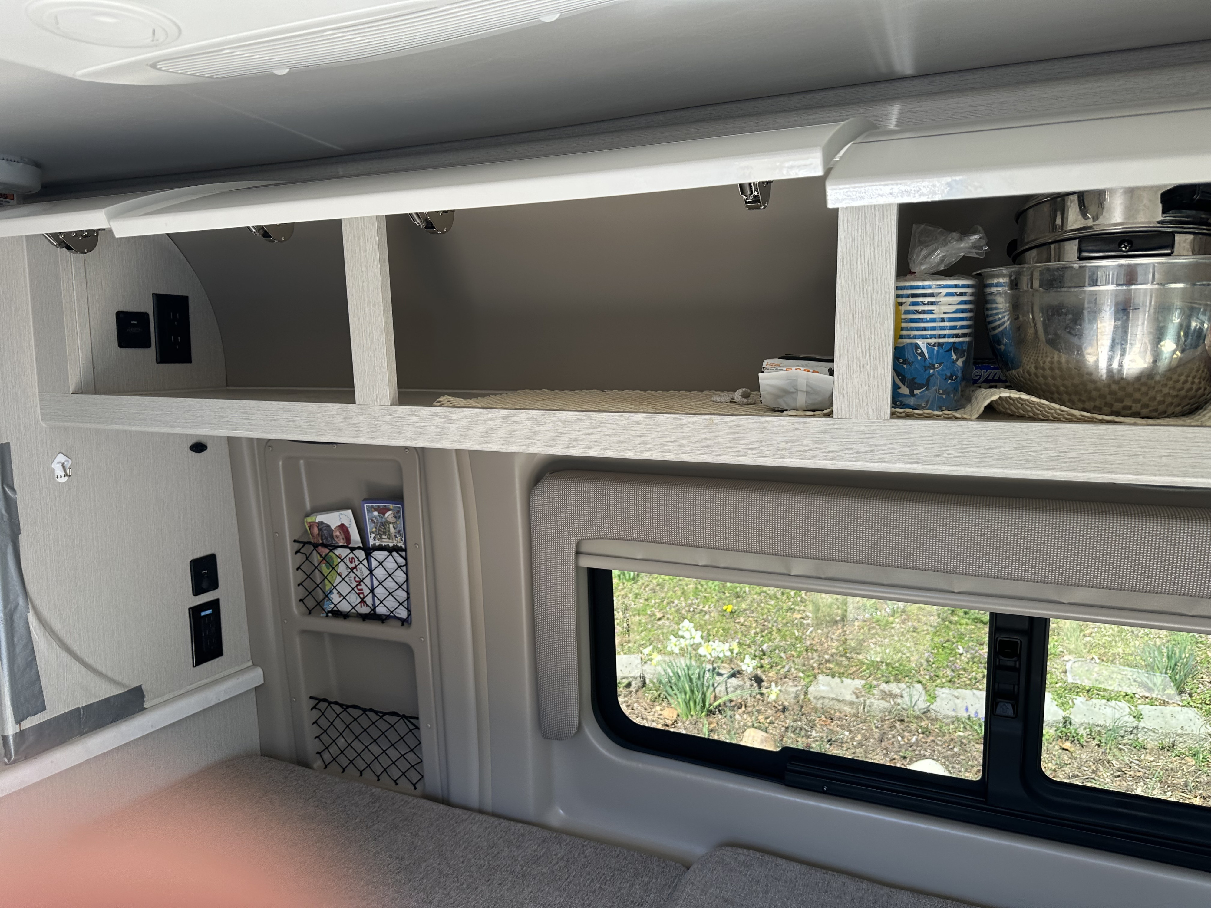 RV rental interior featuring open overhead cabinets with kitchenware, wall outlets, storage nets, and a window view to nature.. Thor Rize 2024