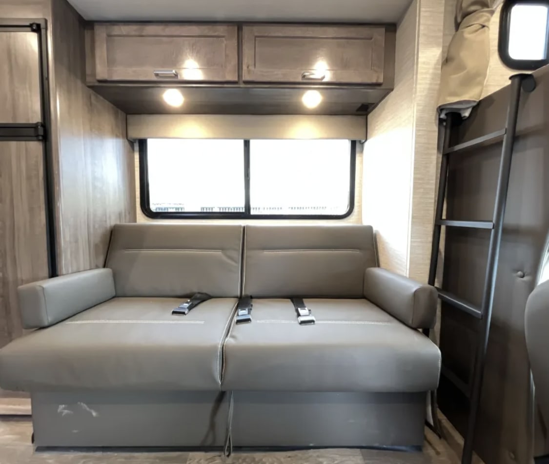 RV rental interior: gray sofa with seatbelts, large window, overhead cabinets, and bunk bed ladder.. Winnebago Minnie Winnie 2025