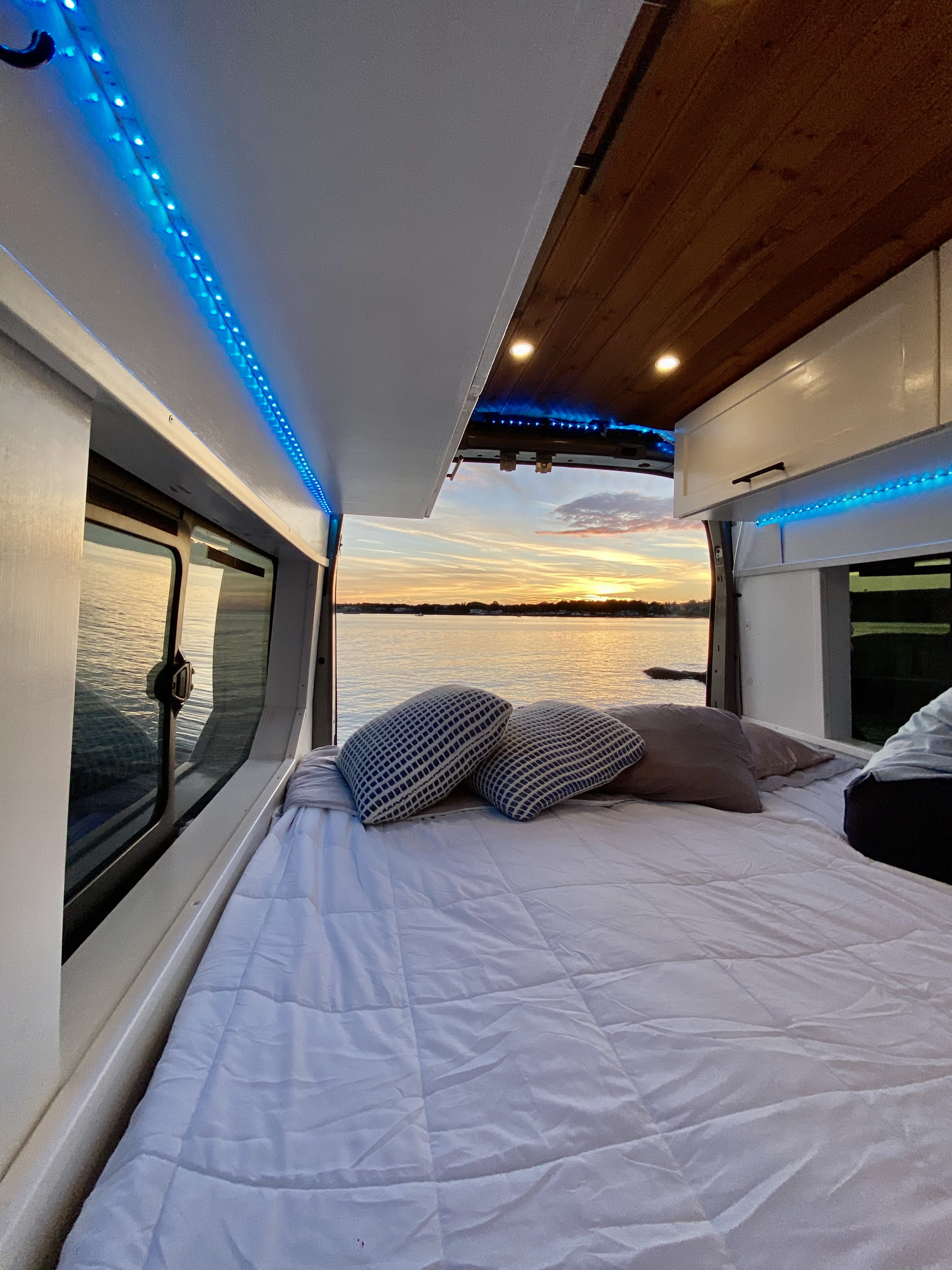 Cozy RV rental interior with bed & pillows, blue lights, and a stunning sunset view over water. Perfect for travel.. Ford Transit 2016