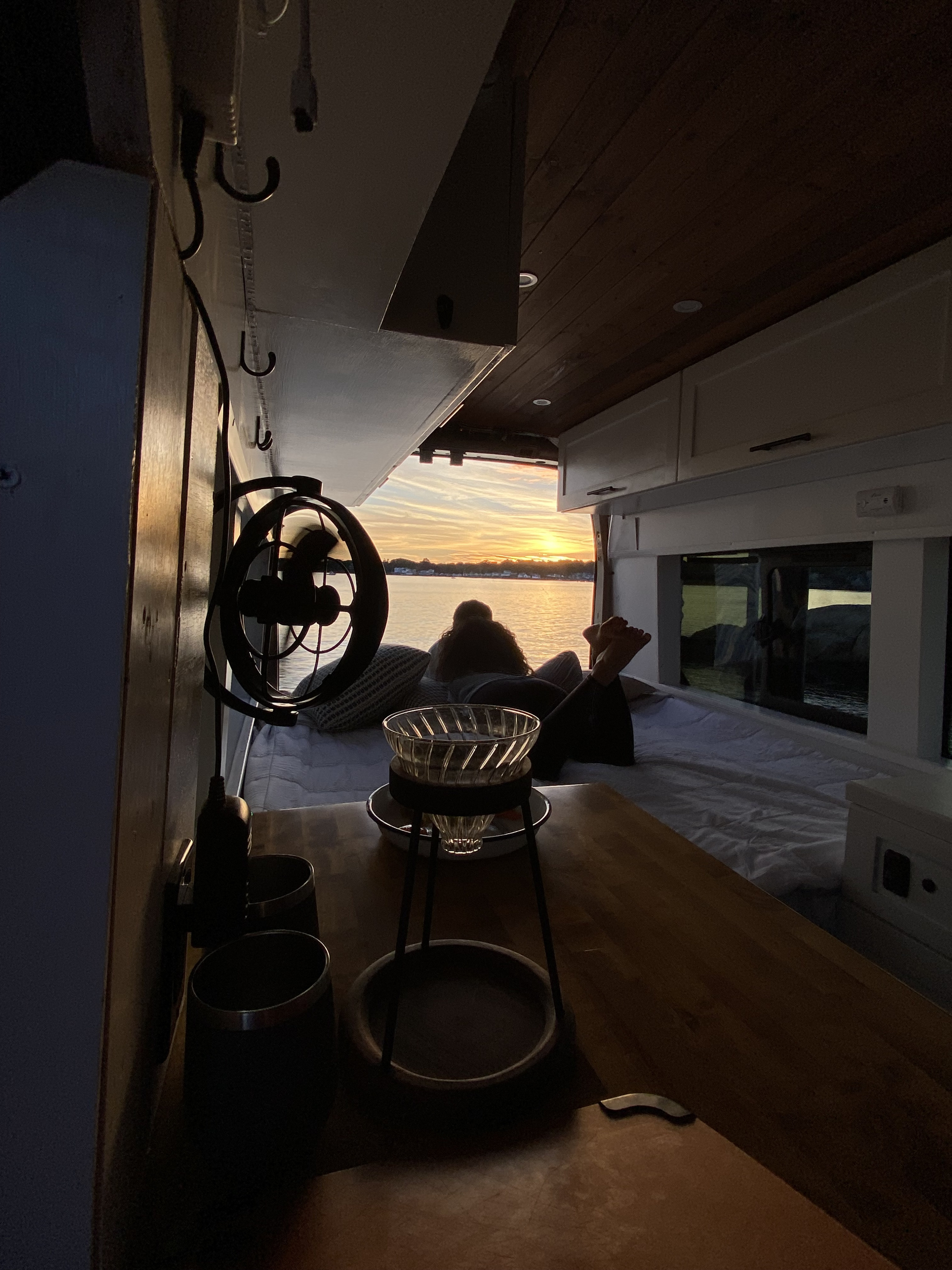 Scenic sunset from a comfy RV rental. A person relaxes on the bed, enjoying the vibrant view over water from inside the camper van.. Ford Transit 2016