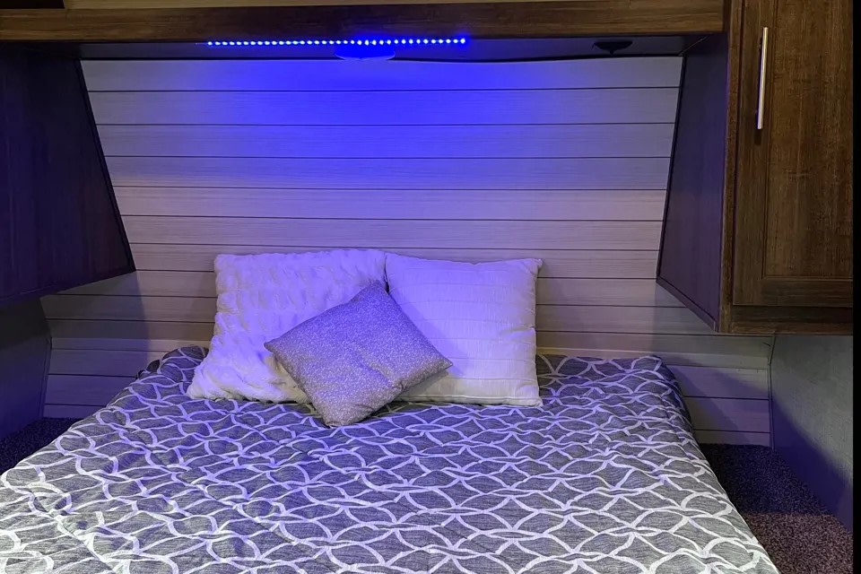 Cozy RV rental bedroom with blue LED lighting, white/grey bedding, light paneled headboard, and wood accents.. Crossroads RV zinger 2019