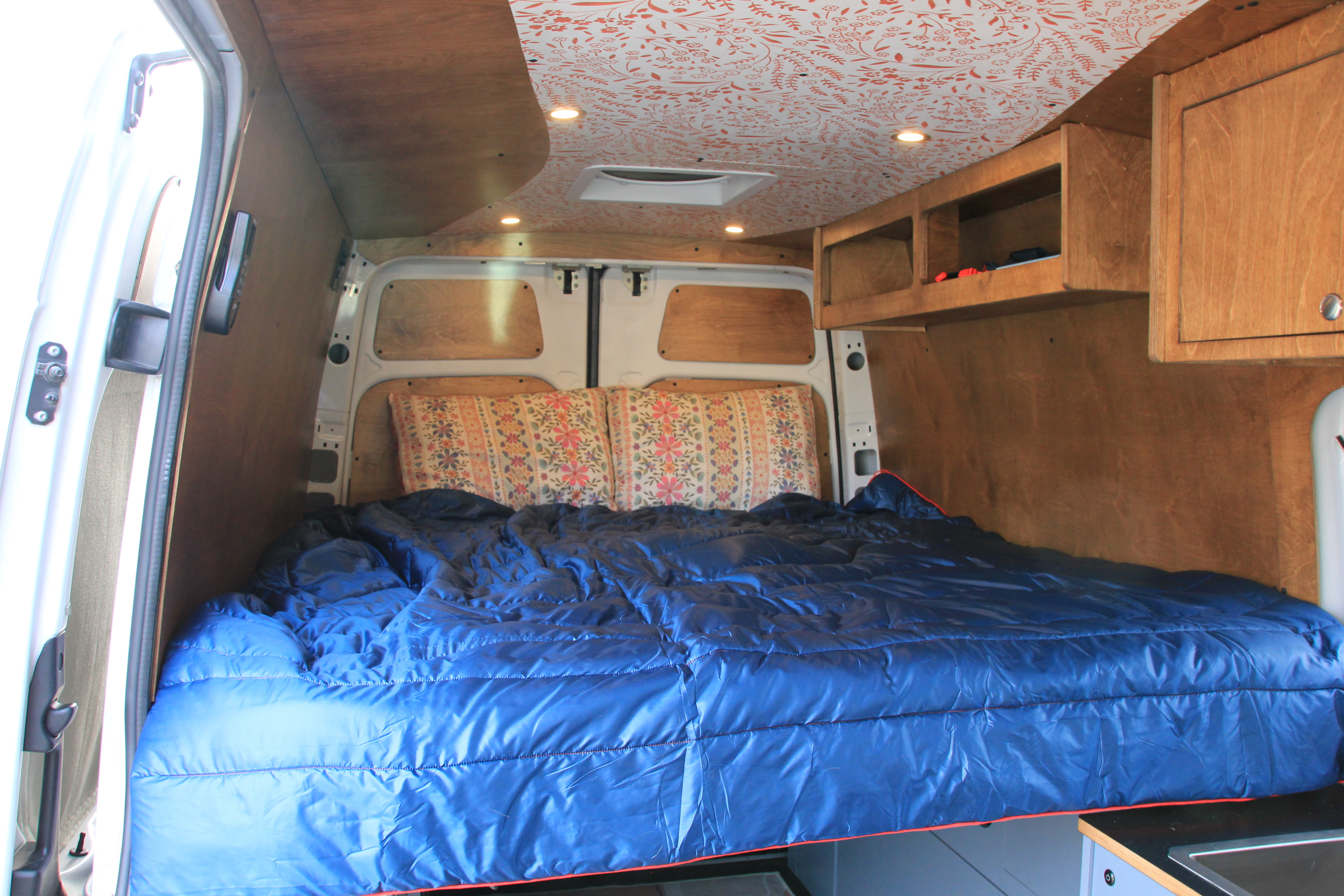 Inside an RV rental: Custom campervan interior featuring a bed with blue bedding, wood walls, cabinets, and a patterned ceiling.. Mercedes-Benz Sprinter 2018
