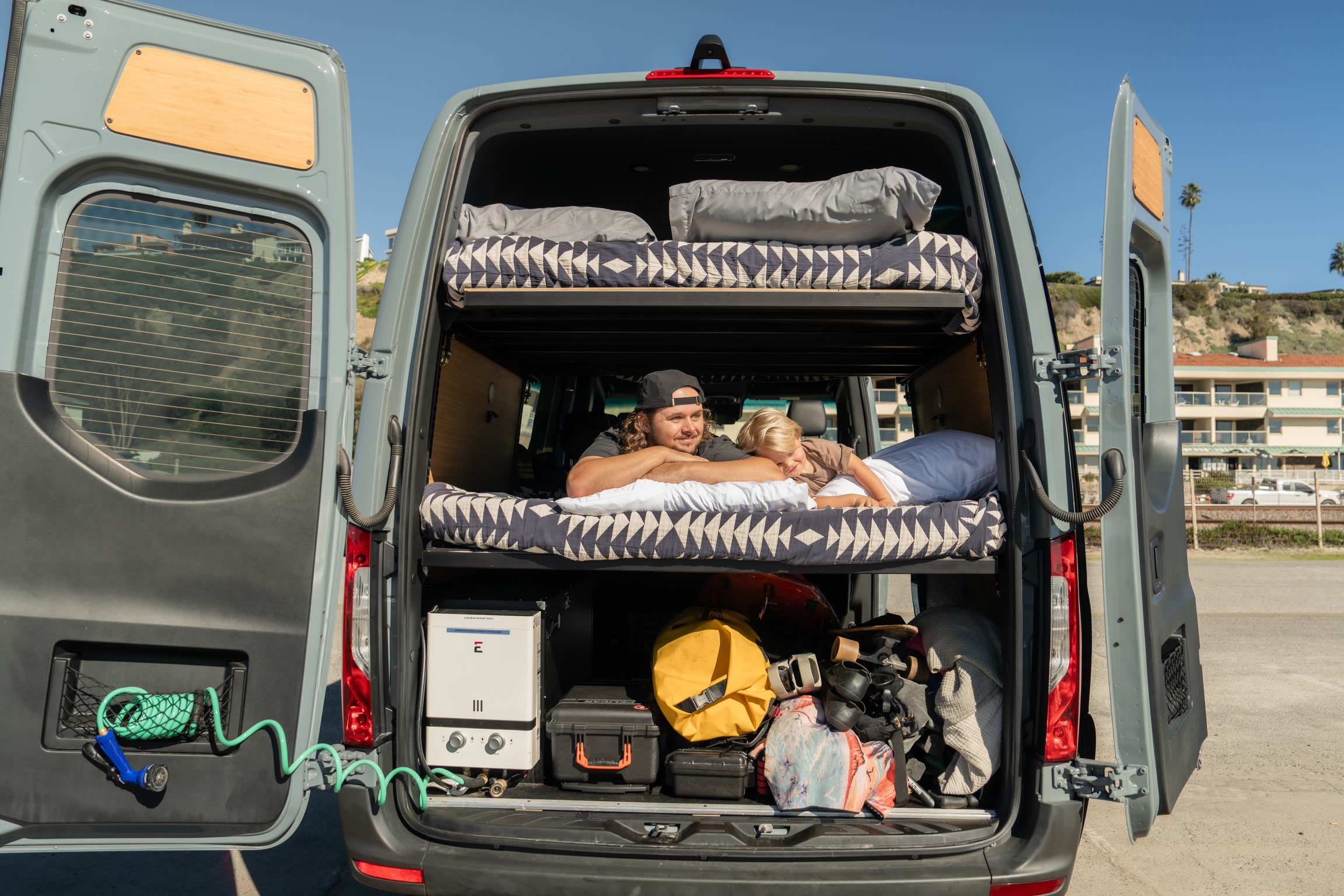 RV rental's open back reveals bunk beds with a man & child, gear storage, and a coiled hose for adventure.. Mercedes sprinter 2025