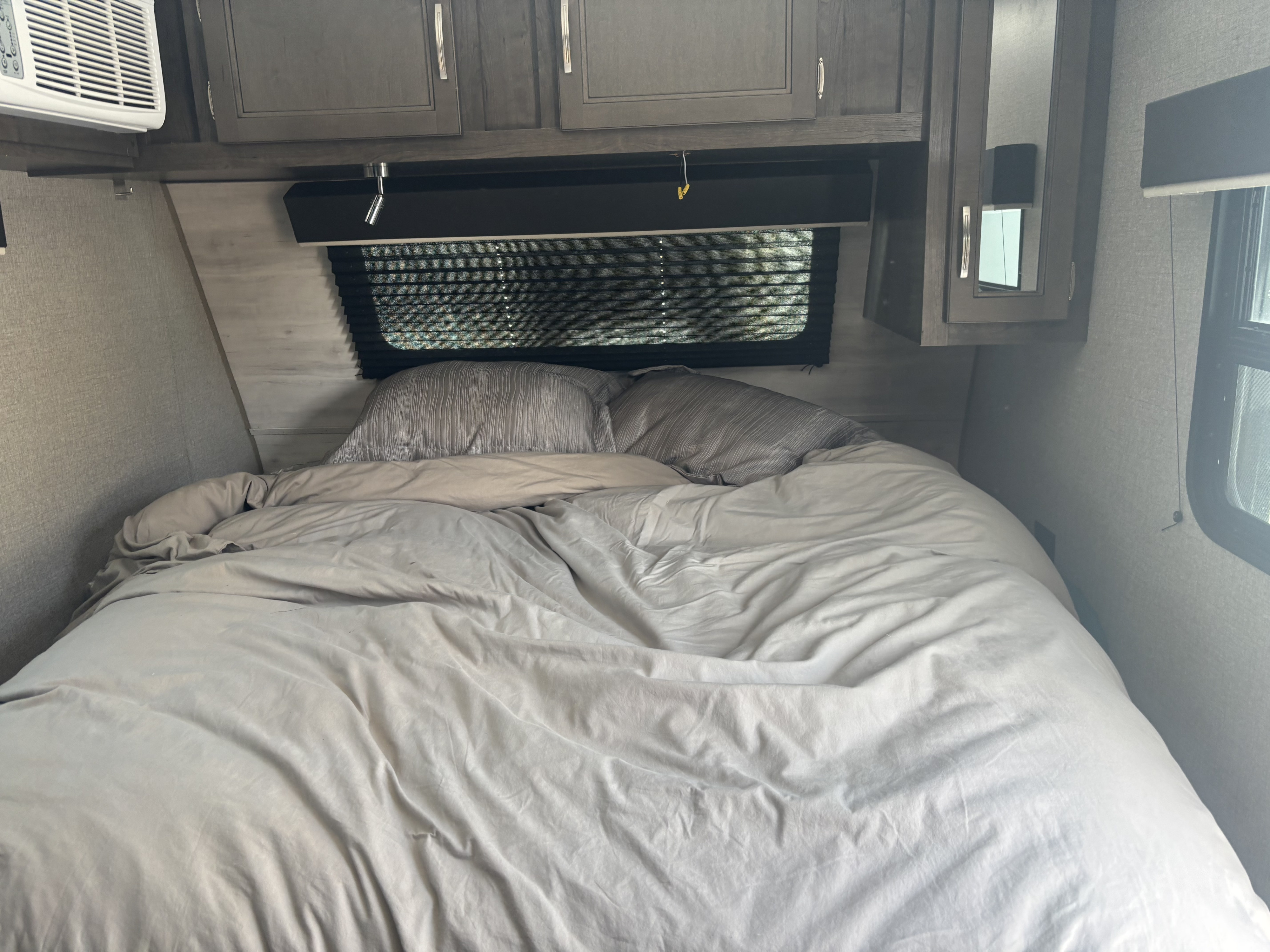 An RV rental bedroom featuring a made bed, gray pillows, overhead cabinets, and a window with blinds, creating a cozy sleeping space.. Jayco Jay Flight SLX Baja Edition 2021