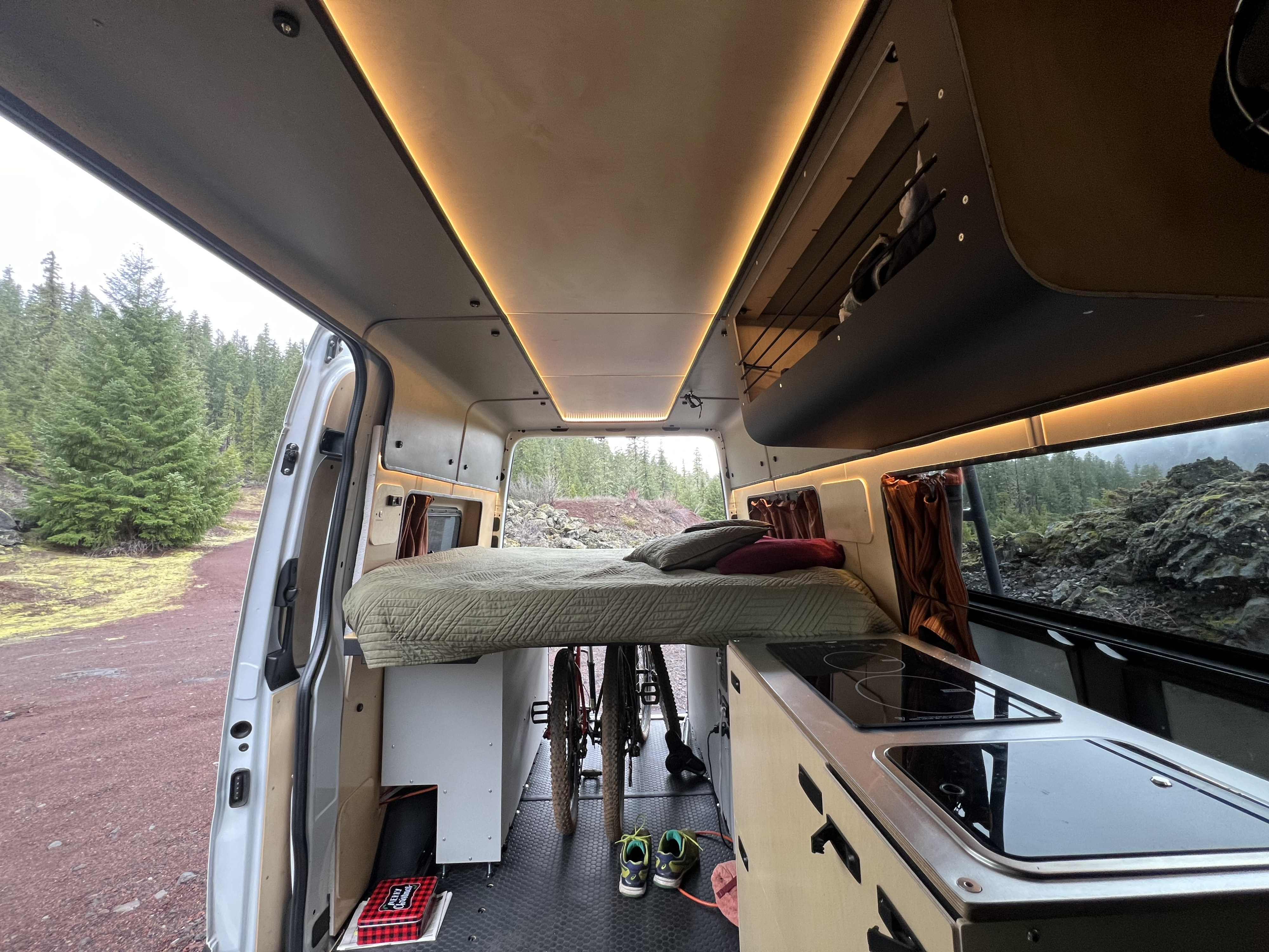 Inside a cozy RV rental: bed, kitchen, bikes below, with a scenic view of evergreen trees & rocks through the open rear.. Mercedes-Benz Sprinter 2021