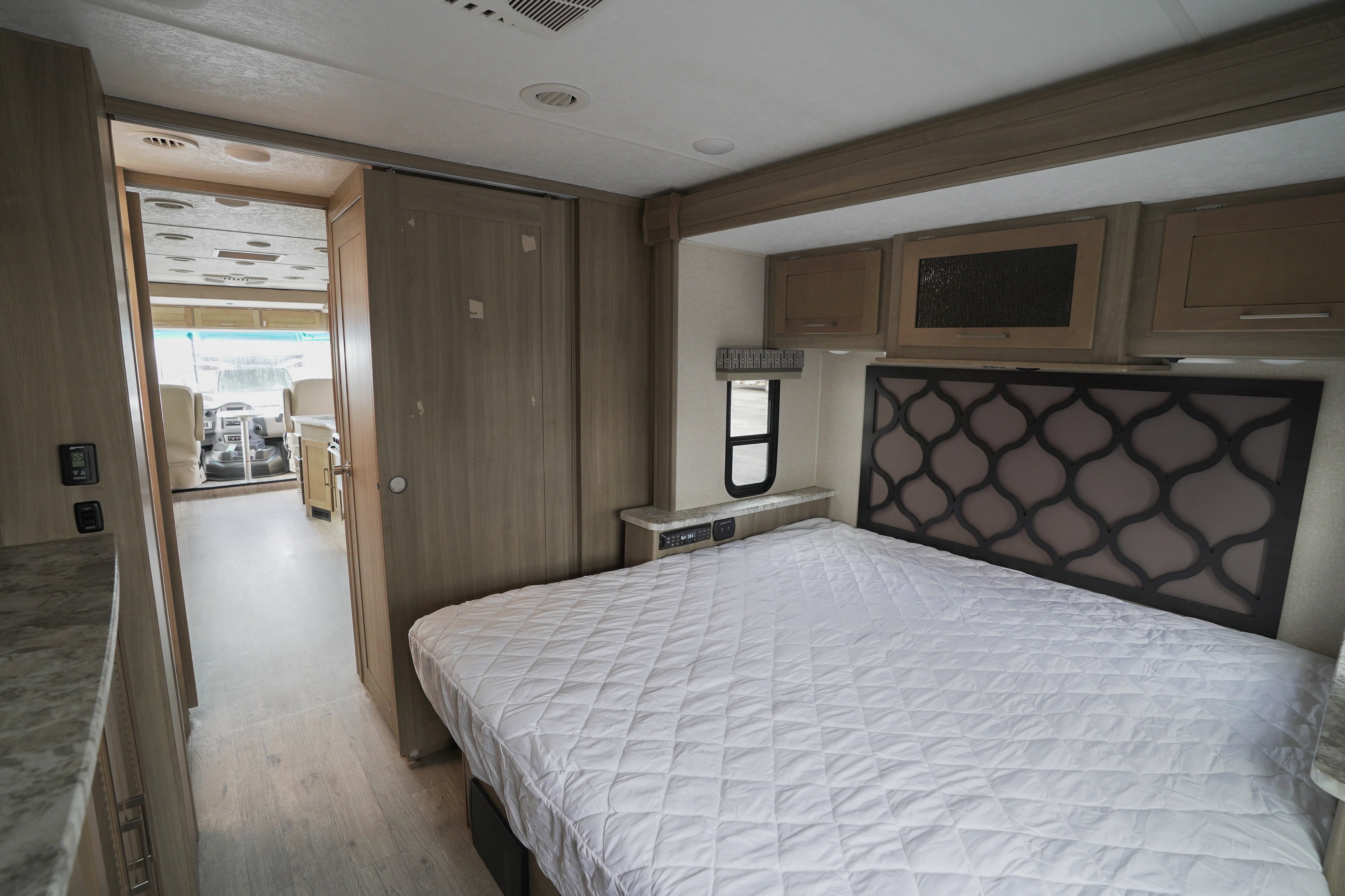 Bedroom view inside an RV rental, featuring a patterned headboard, overhead storage, and access to the main living area.. Coachmen Pursuit 2019