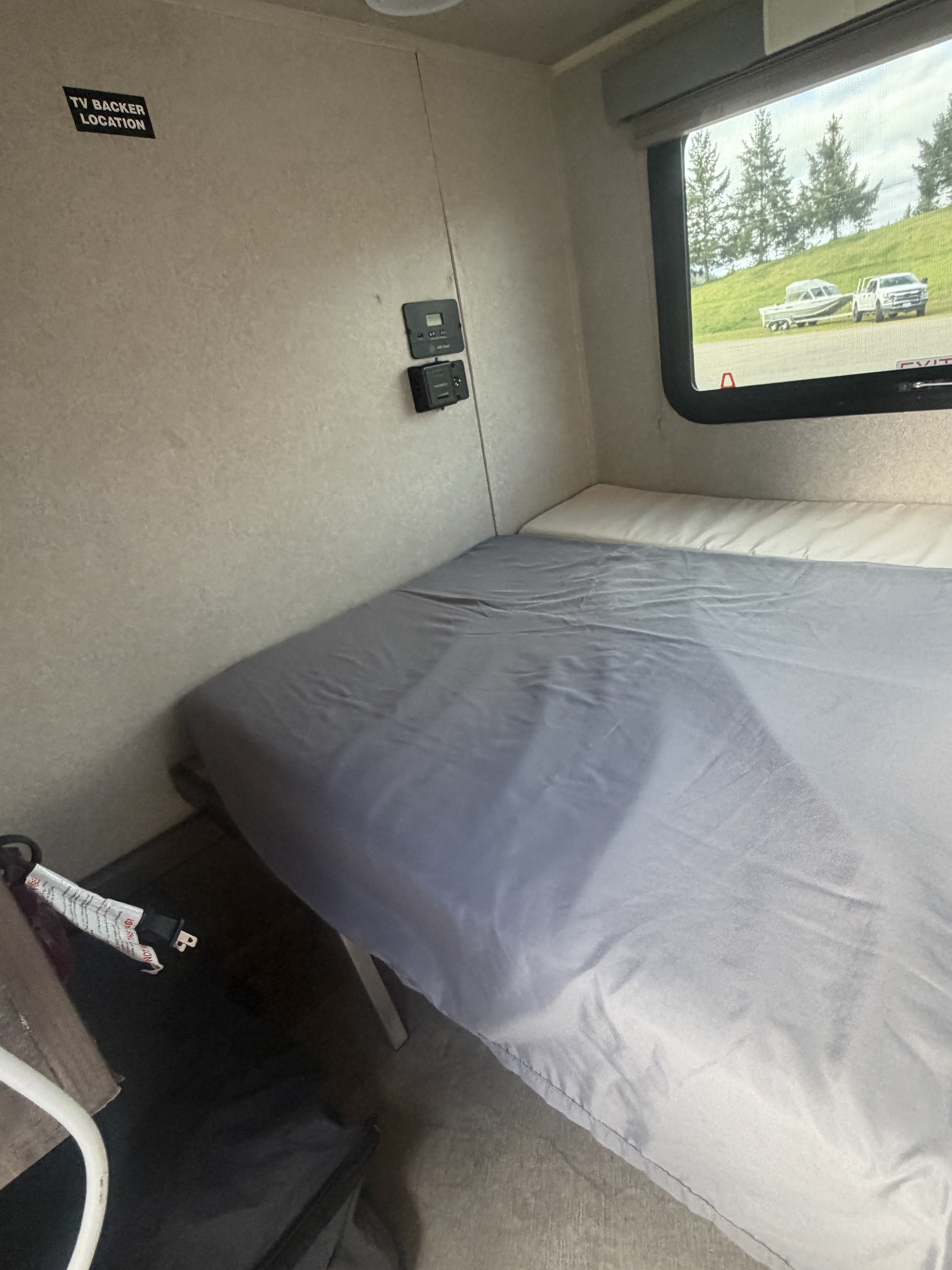 RV rental bedroom with a made bed, wall-mounted controls, and a window view showing a truck, boat, and trees.. Sunset Park RV SunRay 2022
