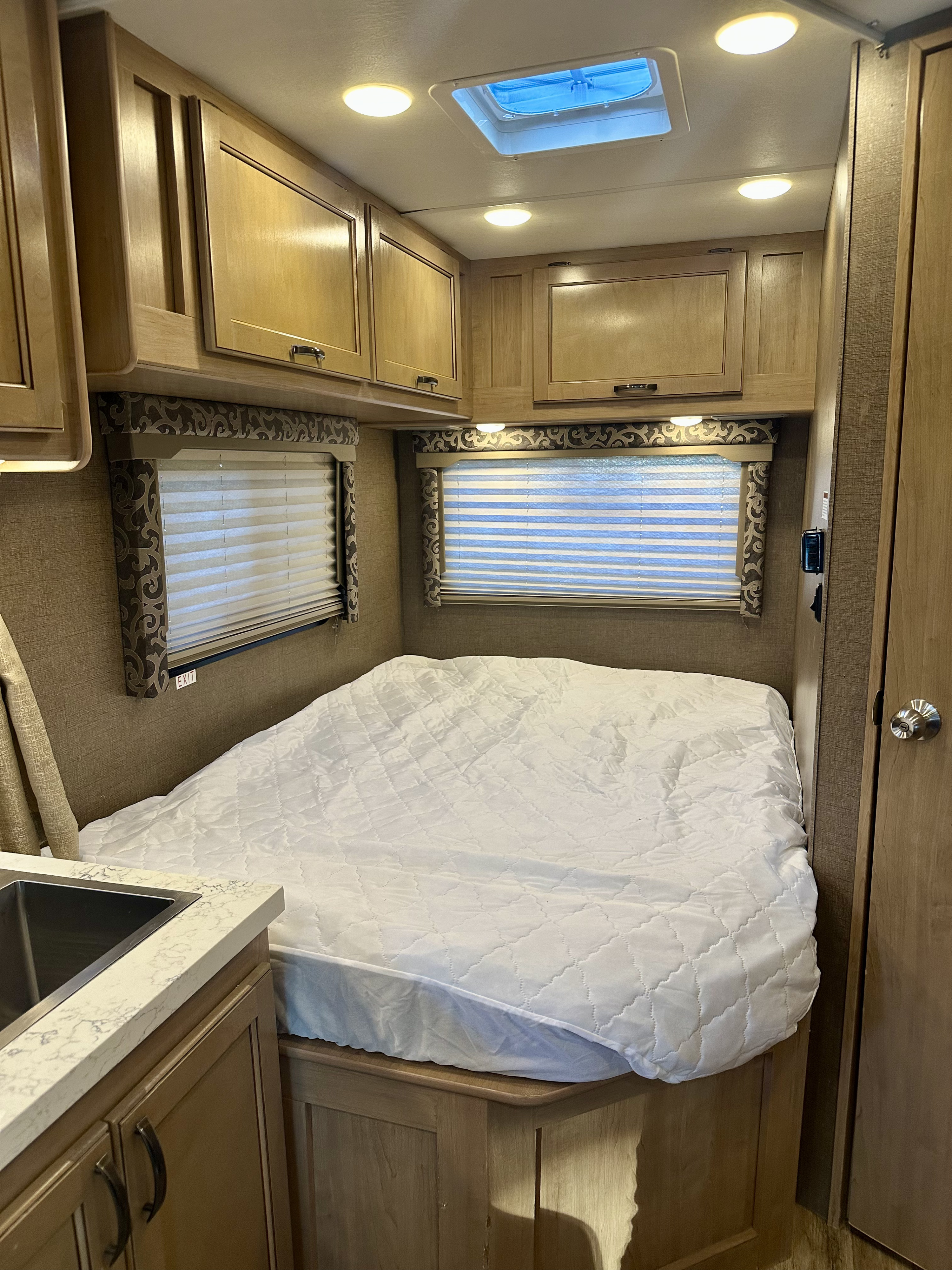 Cozy rv rental bedroom: features a quilted bed, wooden cabinets, windows with blinds, a skylight, and a small counter with a sink.. Thor Four Winds 2019