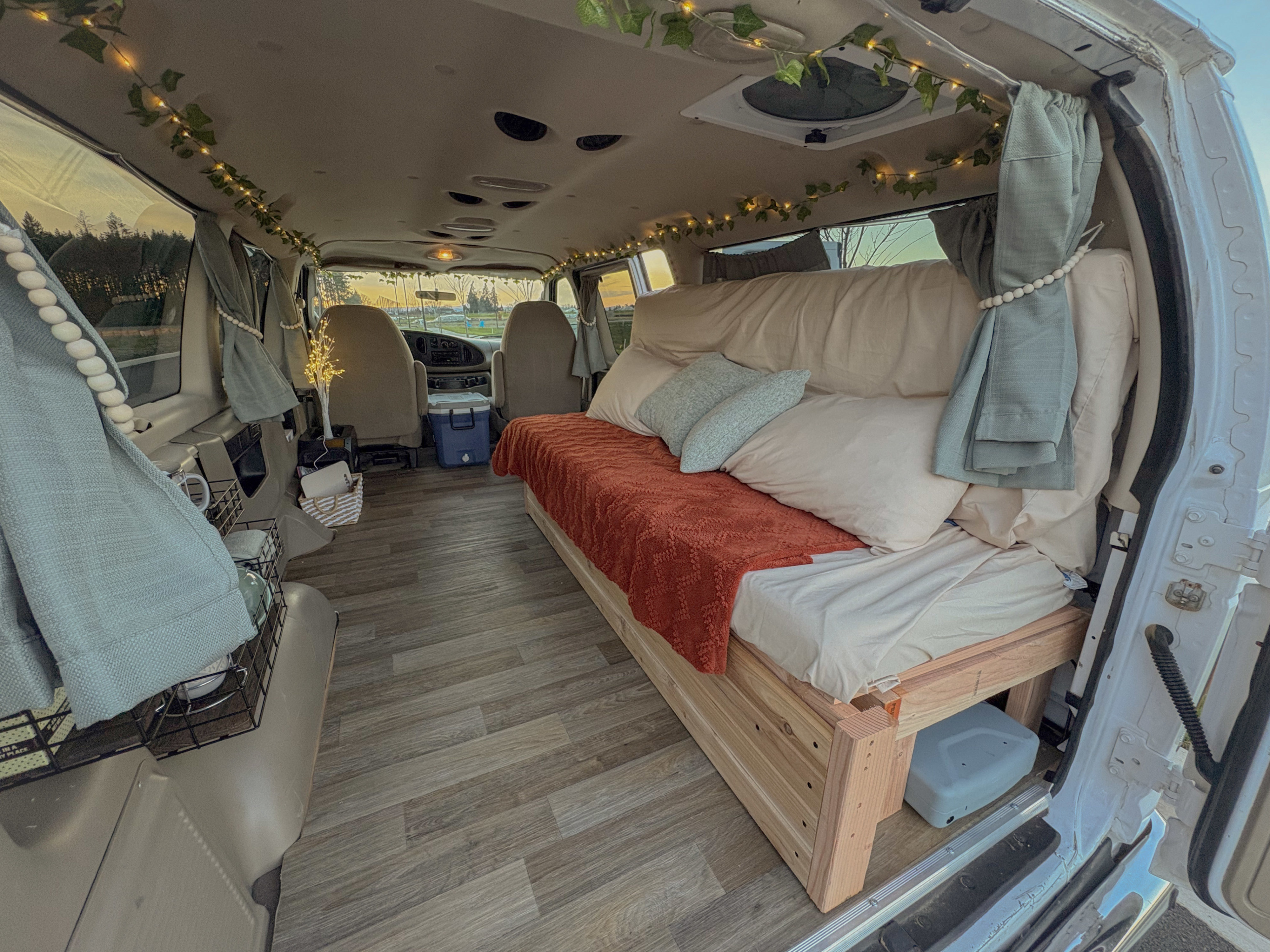 Inside a charming RV rental camper van: a cozy bed with an orange blanket, wooden floor, and ambient fairy lights.. Ford Econoline 2008