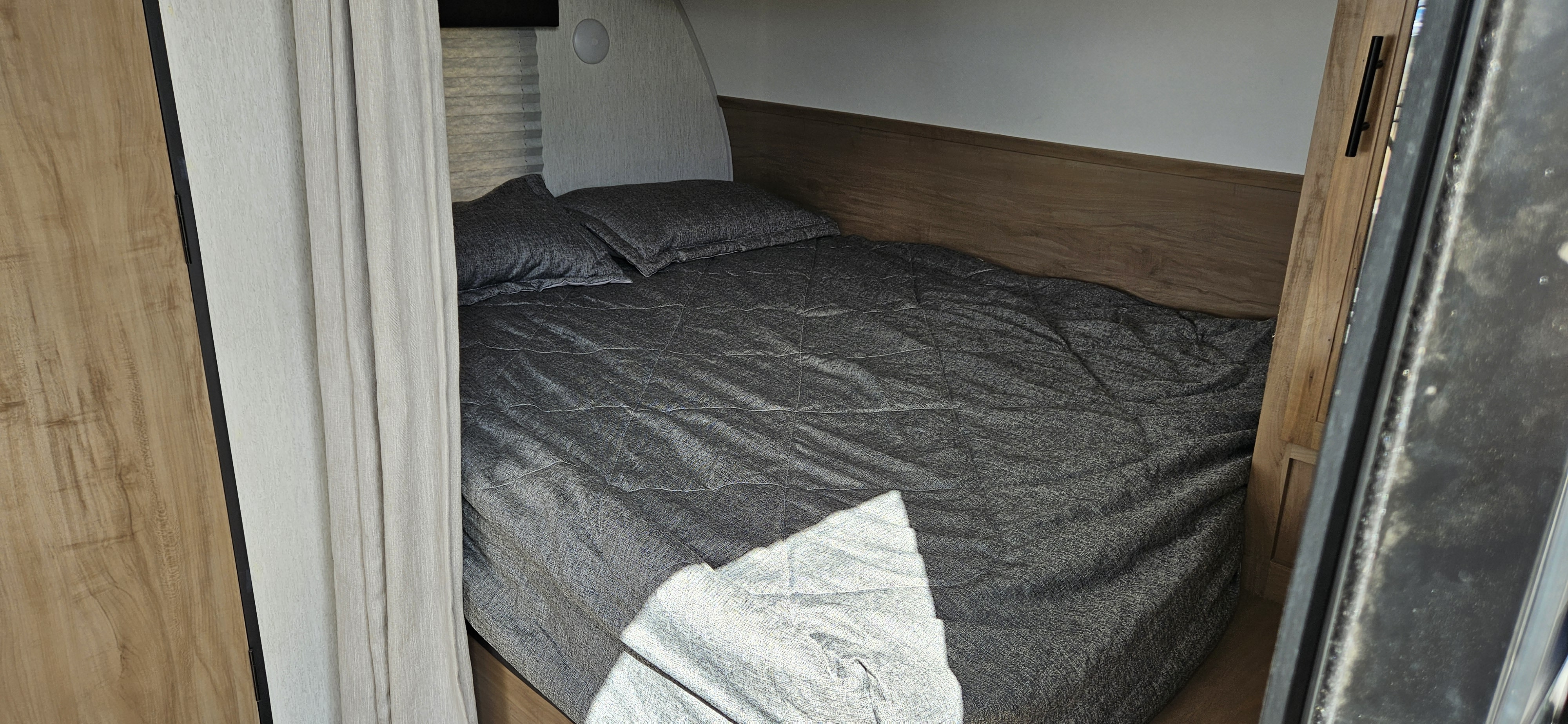 Compact RV rental bedroom with gray quilted bed and pillows, wooden headboard, and curtains, offering a cozy sleeping area.. Forest River Cherokee Grey Wolf 2023