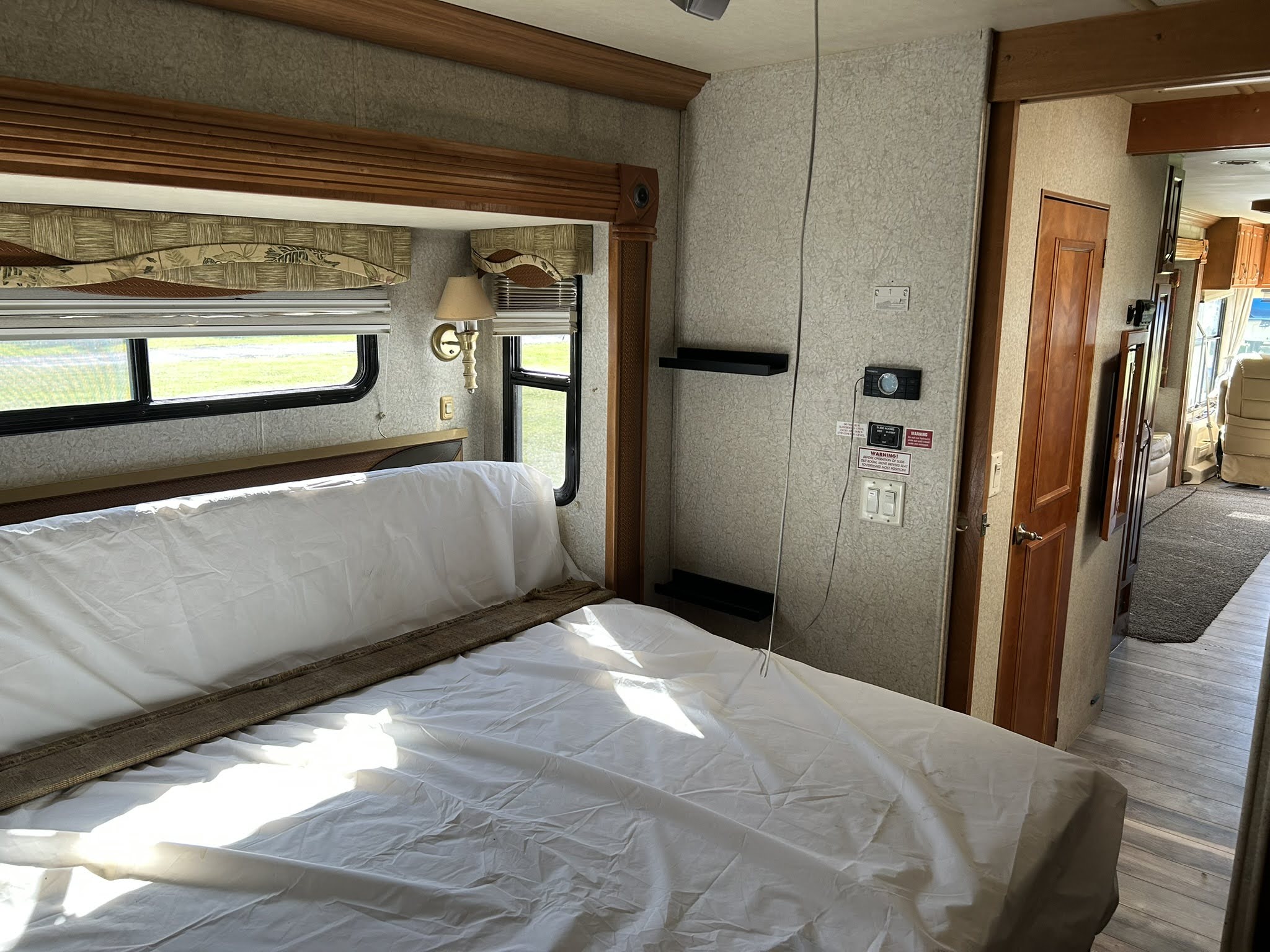 Spacious RV rental bedroom featuring a bed with white sheets, window, wooden trim, and controls, leading to the living area.. Gulf Stream Tourmaster 2005